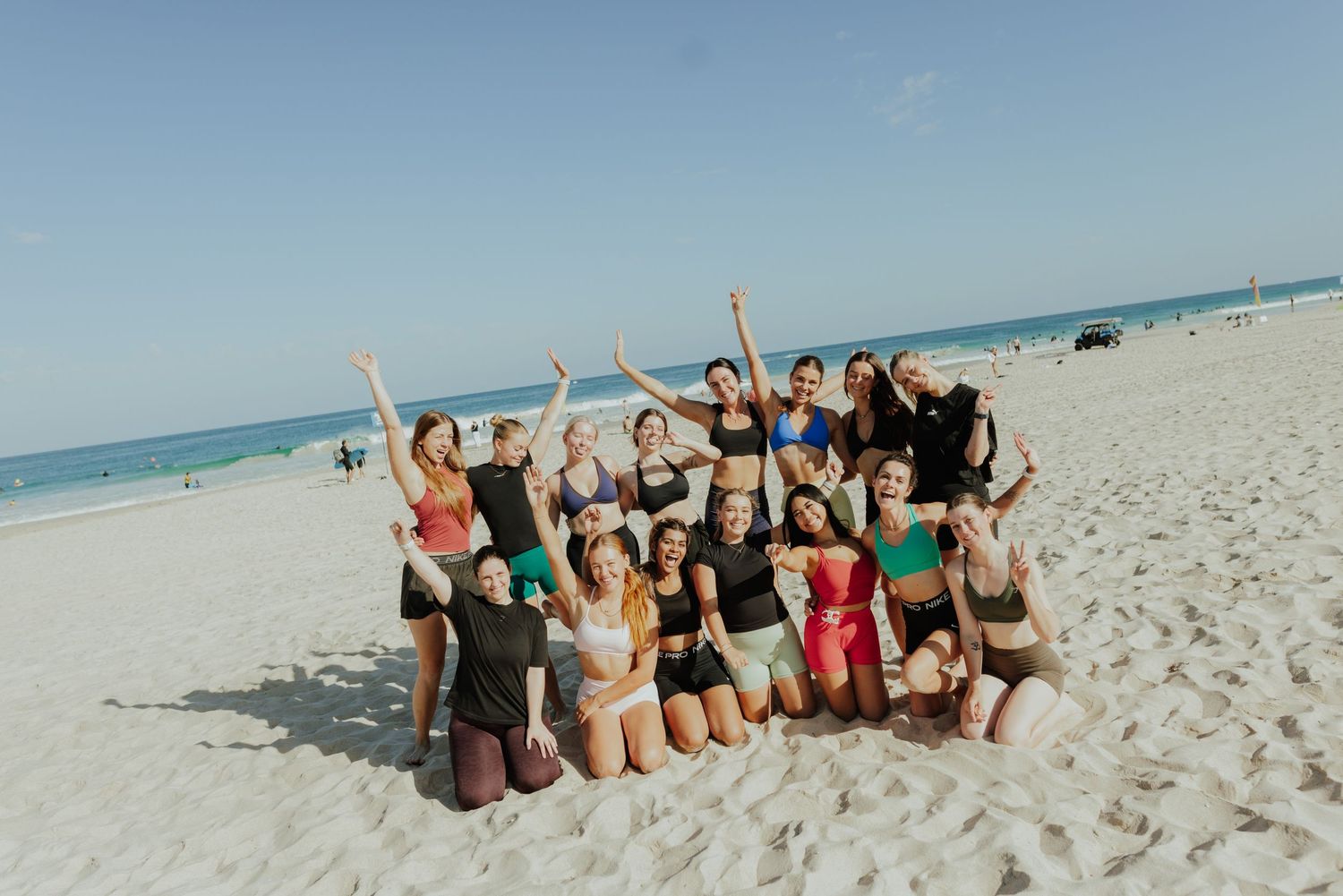 Group of women posing together on a white sandy beach with hands raised in celebration.