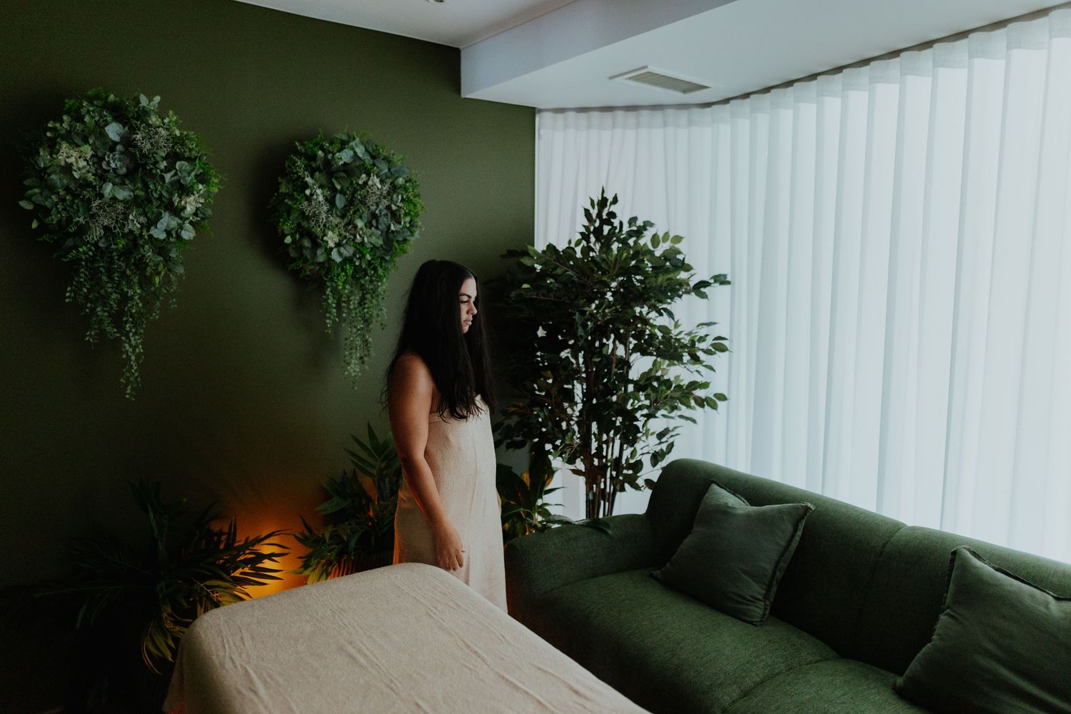 Woman in white dress standing in a serene green-themed spa room with lush plants and floral wall decor.