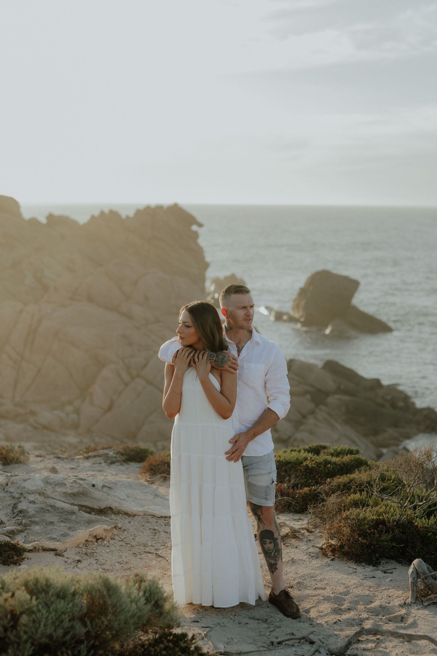 Couple embracing on rocky coastal cliff at golden hour with ocean views behind them.