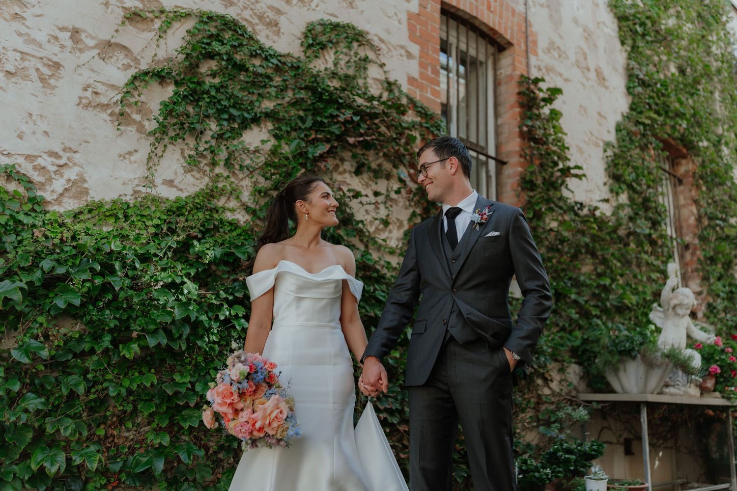 Bride in white mermaid gown and groom in grey suit hold hands before an ivy-covered stone wall.