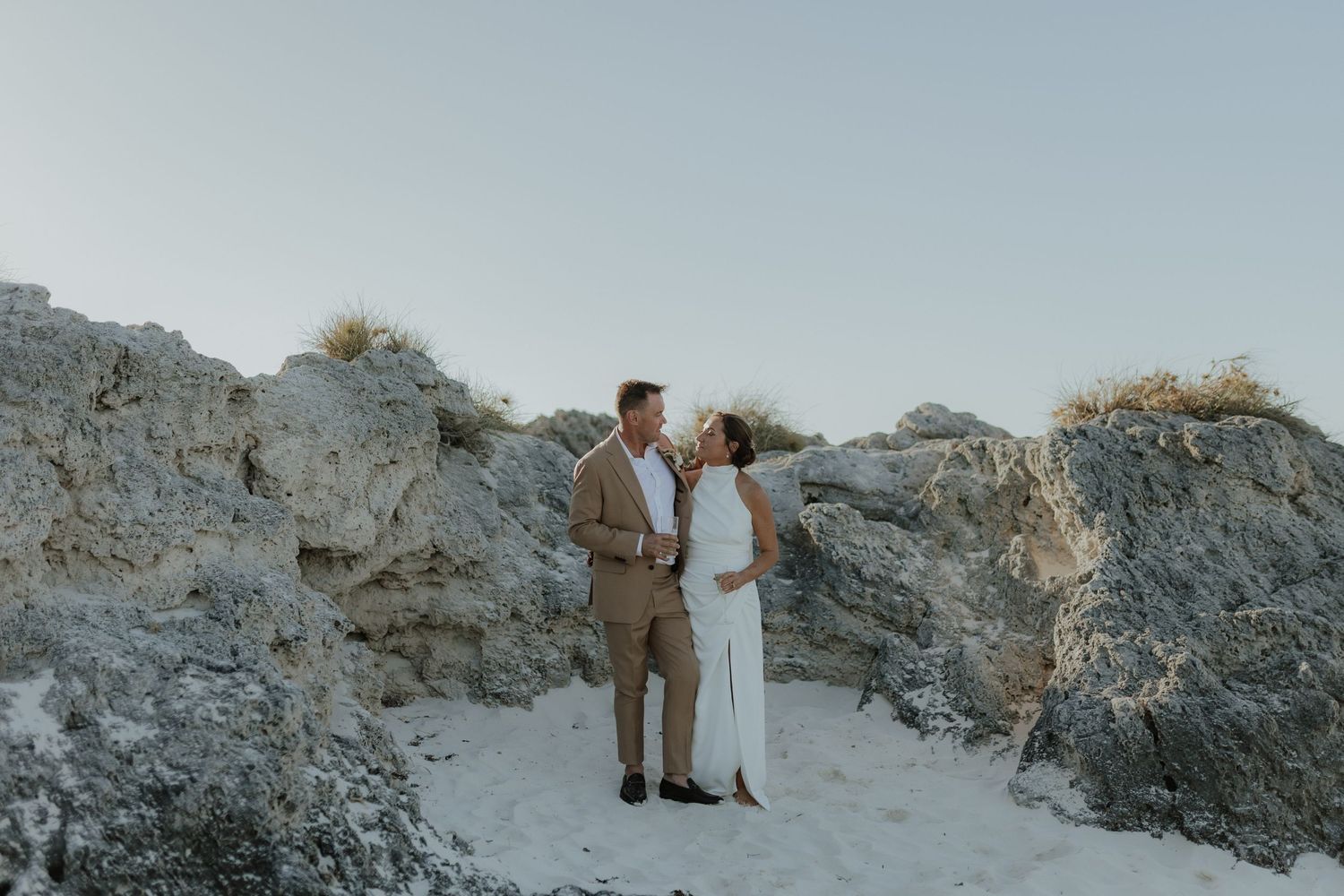 Couple in wedding attire standing among rocky formations on a sandy beach at dusk.