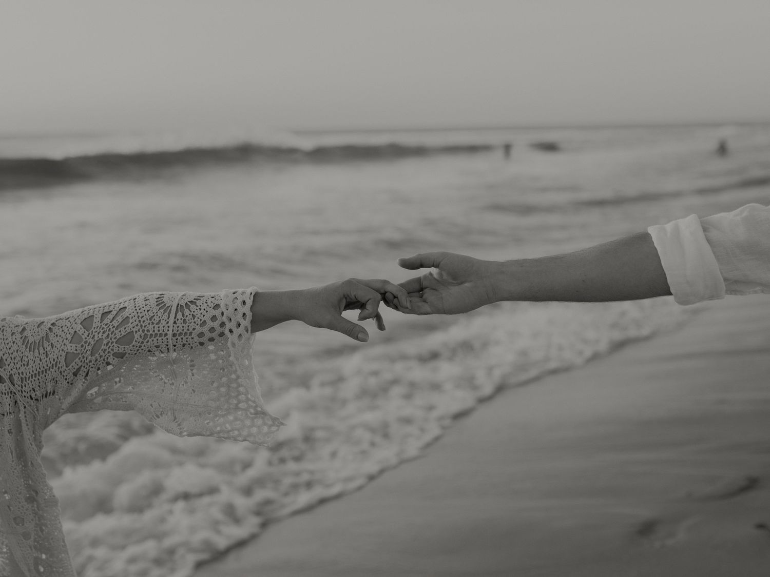 Black and white close-up of couple gently touching fingertips on ocean shore.