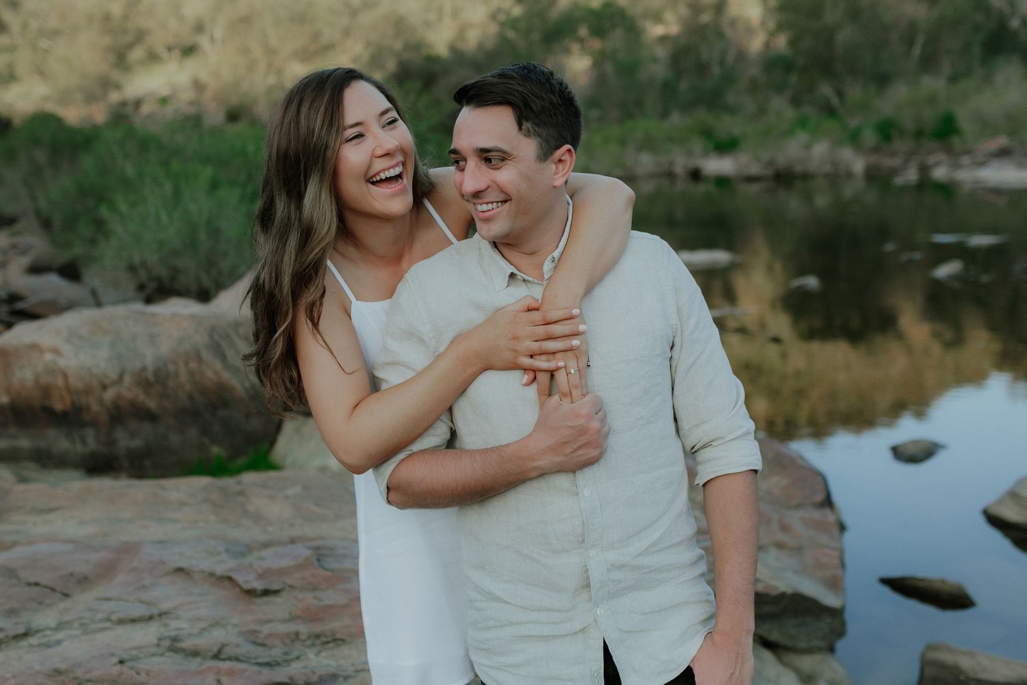 Laughing couple embracing near a tranquil river surrounded by natural rocky scenery.