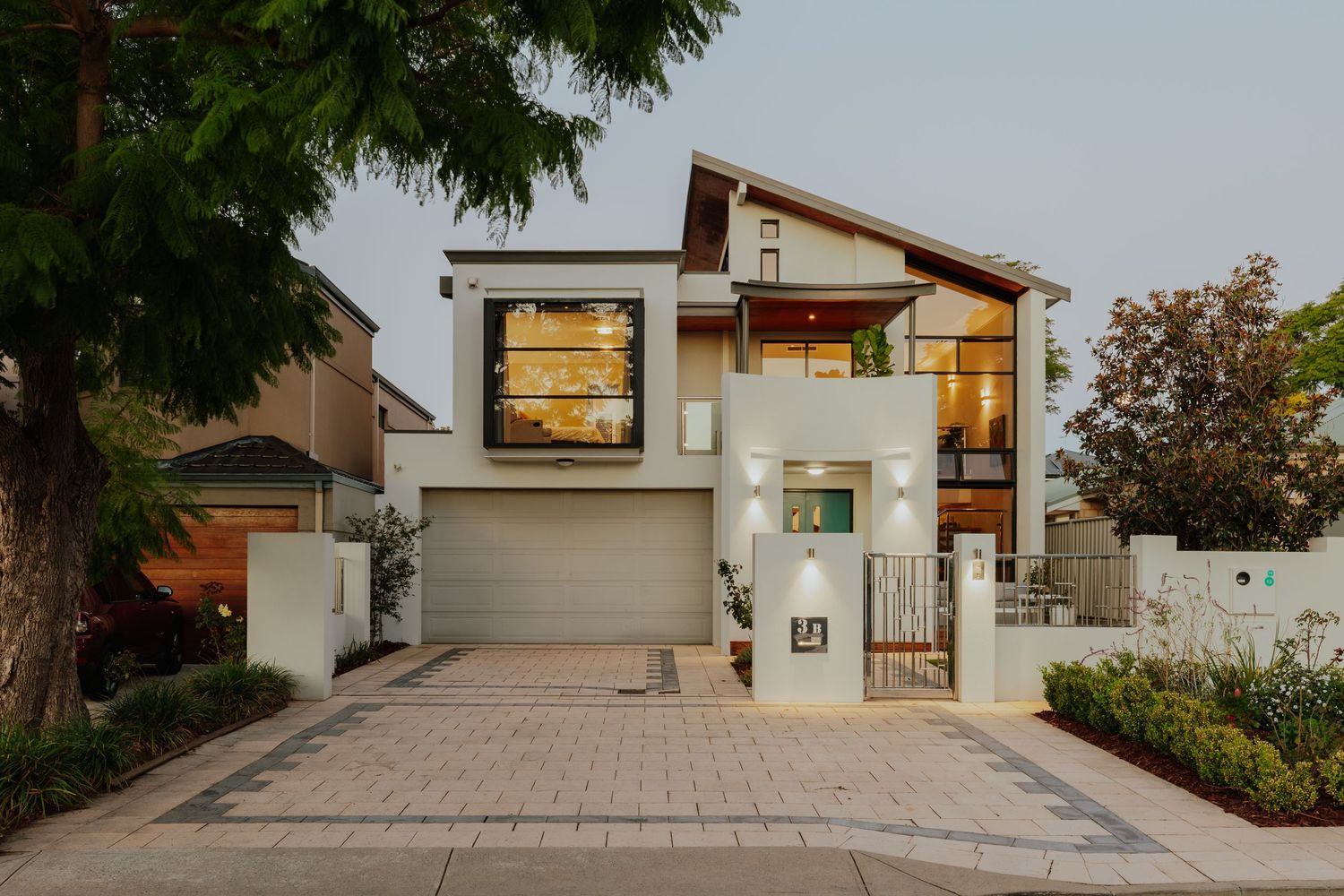 Modern two-story suburban home with white render, timber accents, glass balcony, and paved driveway at dusk.