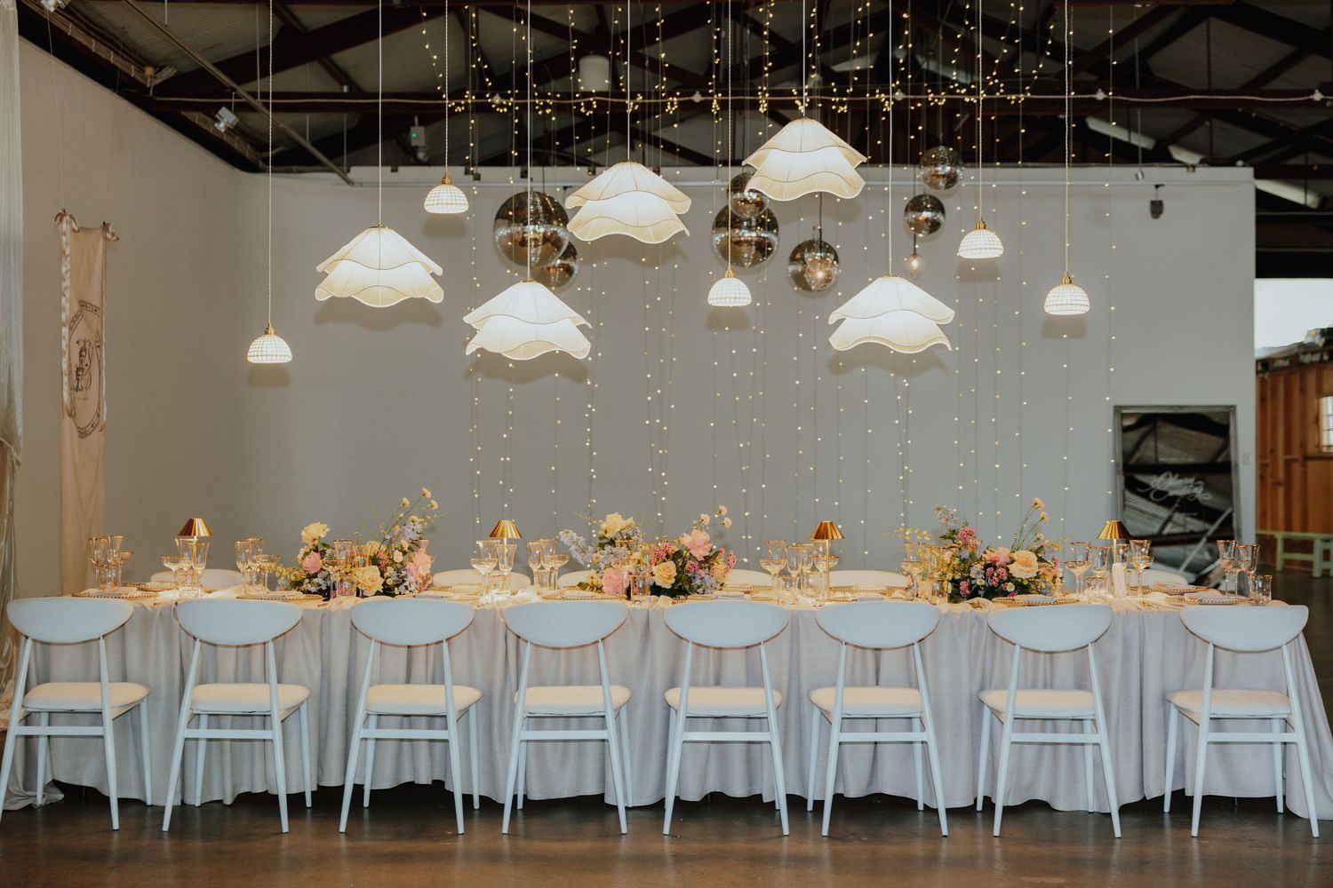 Long wedding reception table with pastel floral centerpieces beneath white cloud-shaped pendant lights.