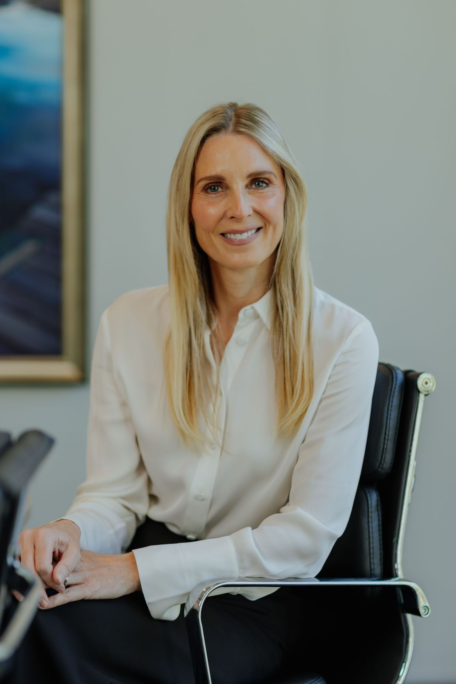 Smiling blonde businesswoman in cream blouse seated in black office chair in professional setting.