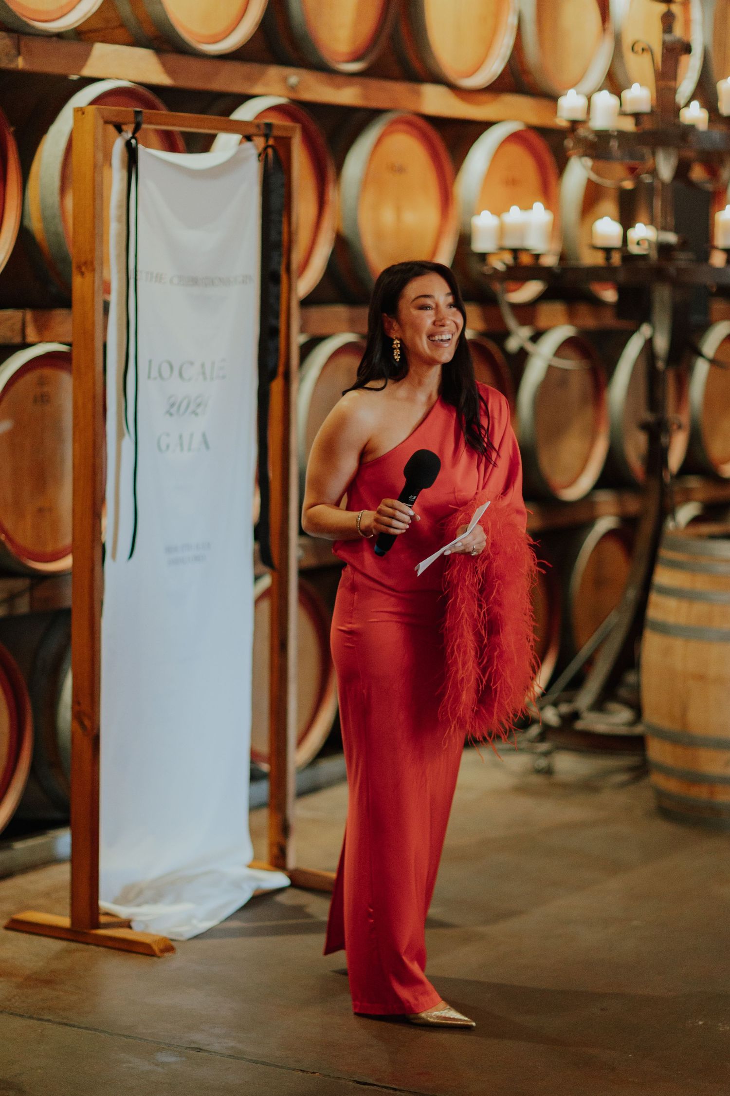 Woman in red feathered jumpsuit hosting a gala event with microphone in a winery barrel room.