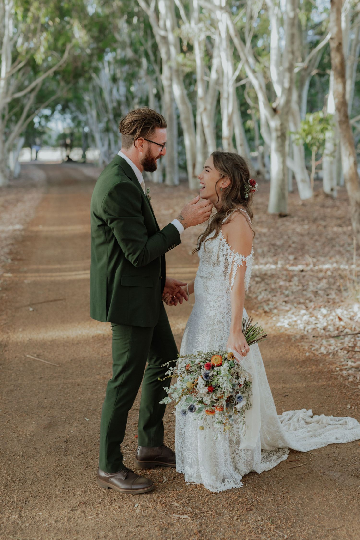 Groom in green suit tenderly touches brides face as they stand together on tree-lined dirt path.