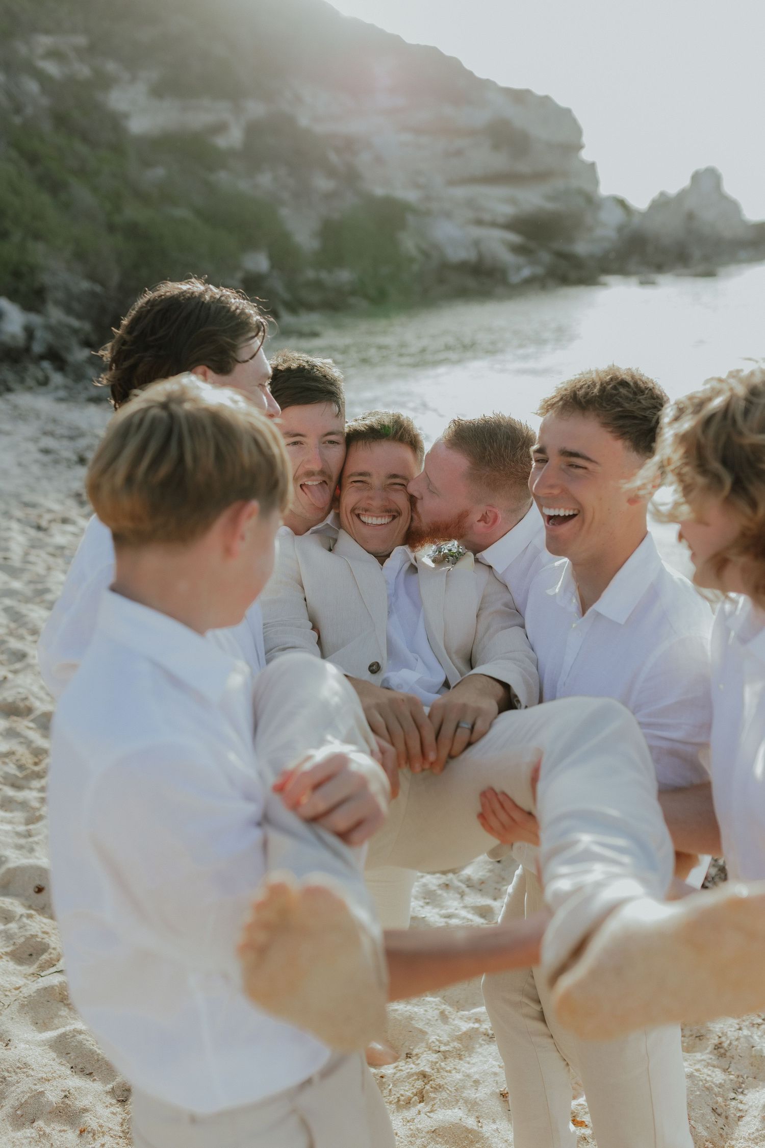 Groom laughing with groomsmen in white shirts sitting together on a sandy beach.