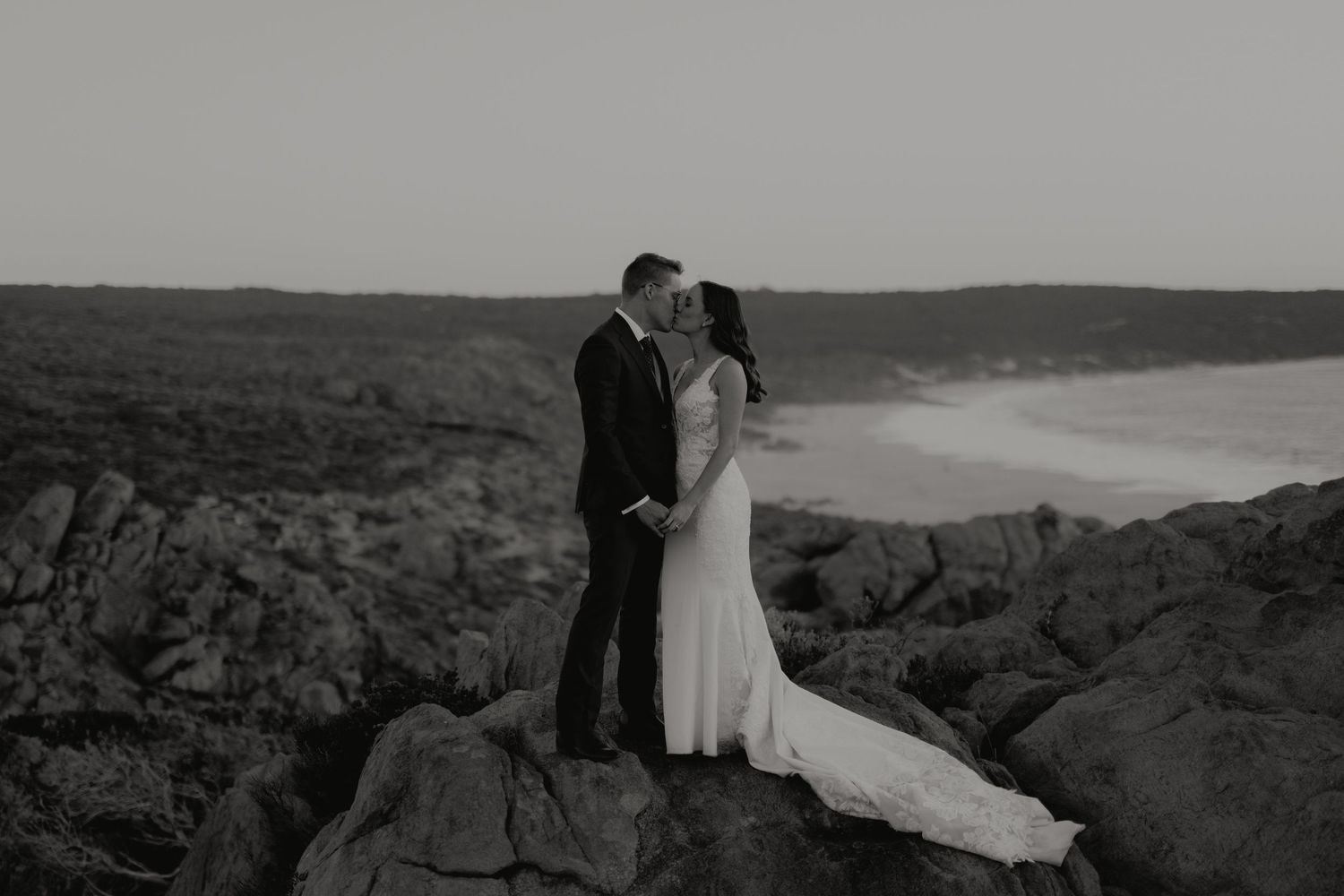 Black and white photo of bride and groom kissing on rocky coastal cliffs at sunset.