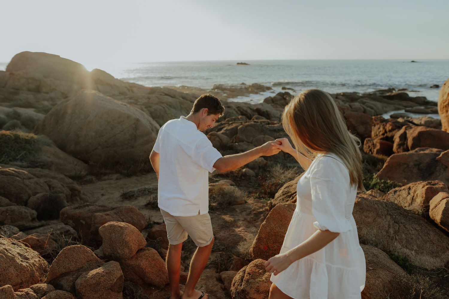Couple in white outfits holding hands while climbing rocky coastal boulders at golden hour sunset.