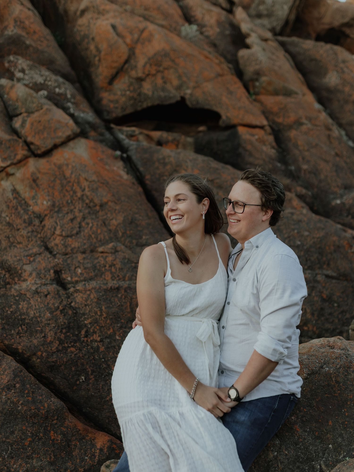 Happy laughing couple sitting against textured red rock face during outdoor engagement photo session.