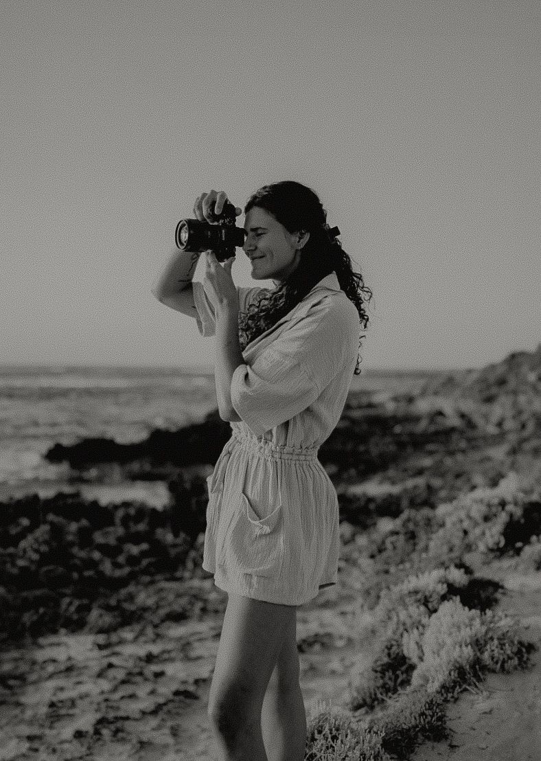Woman in white romper photographing at rocky beach with film camera, black and white photography.