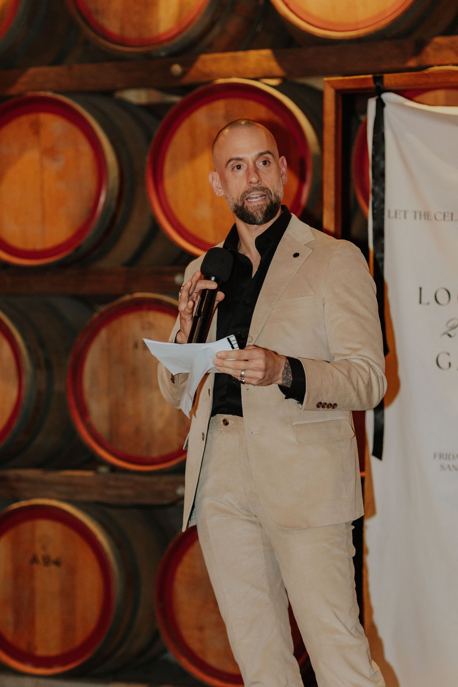Man in beige suit speaking at a podium with notes at a winery gala event.