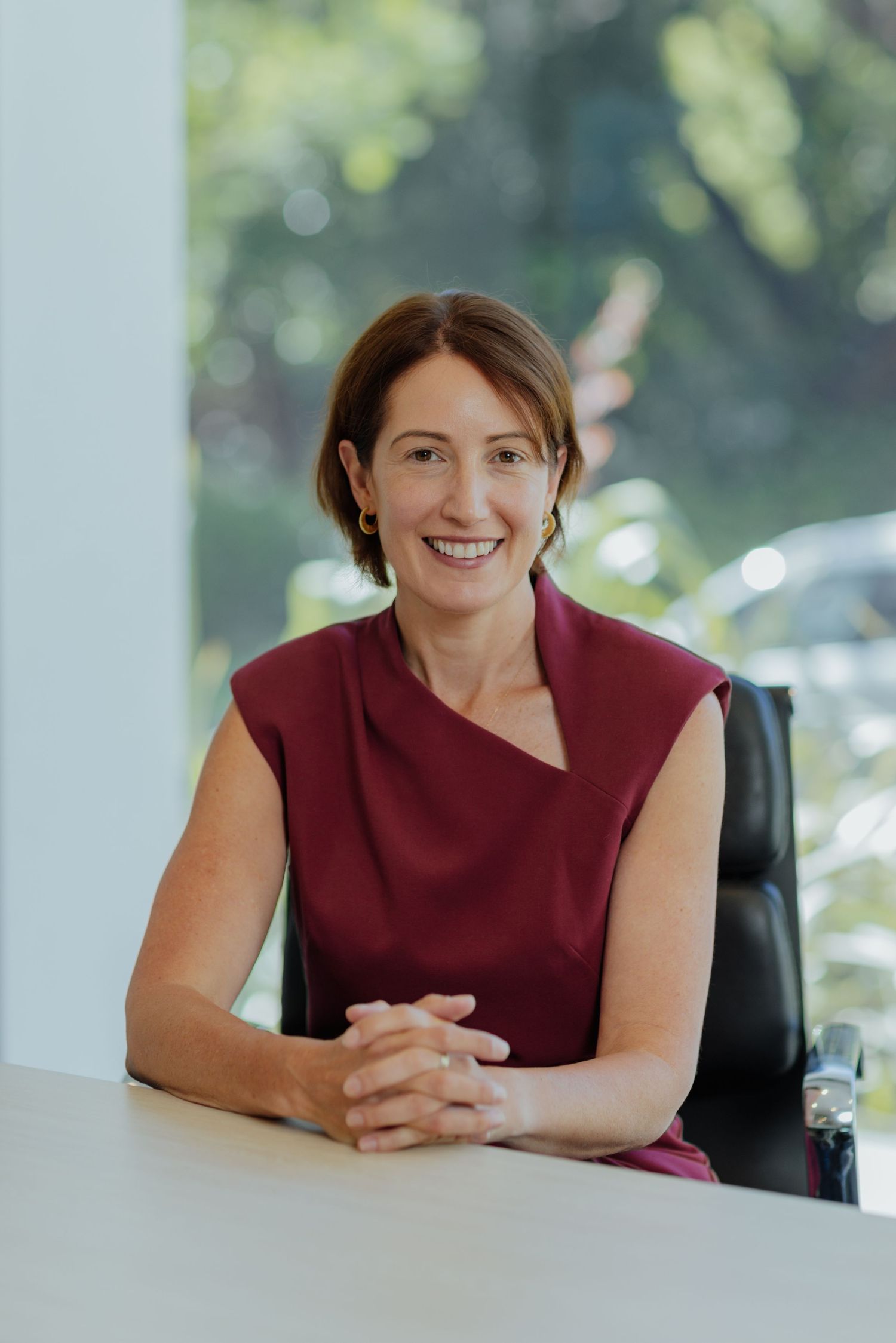 Smiling businesswoman in burgundy dress seated at a white desk in a bright modern office space.