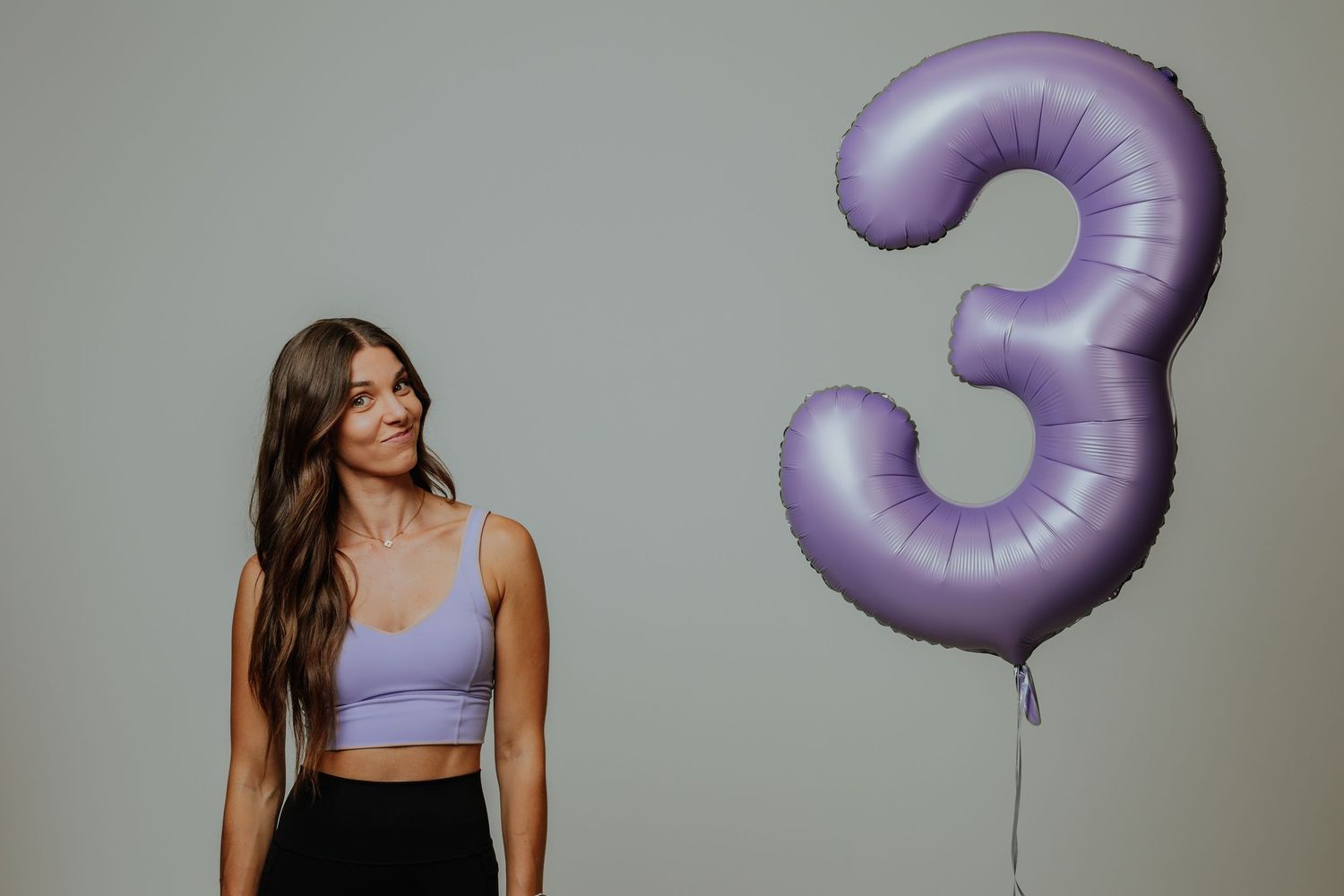 Smiling woman in lavender crop top standing next to a large purple number 3 balloon.