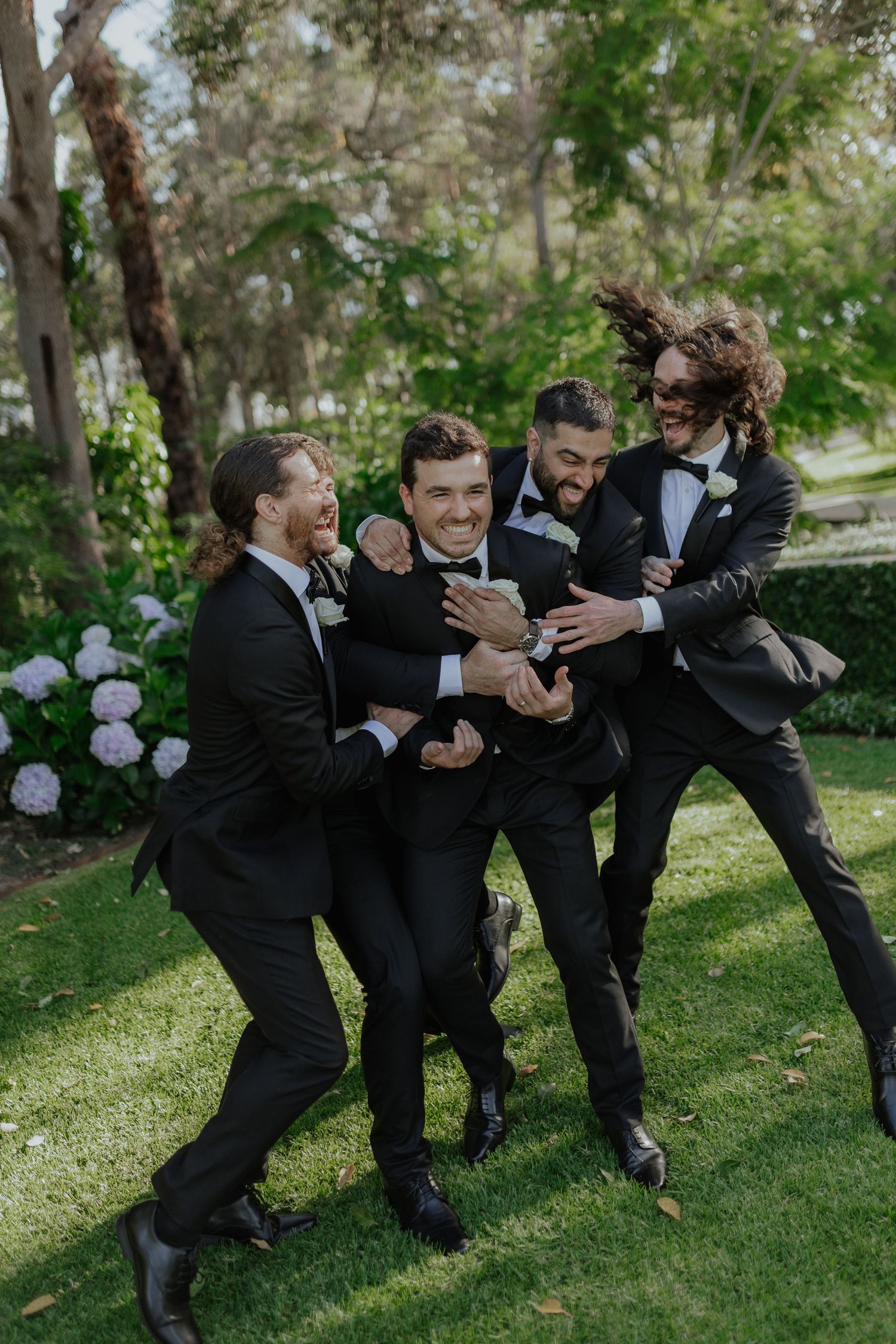Four groomsmen in black tuxedos laugh and celebrate together on a green lawn surrounded by lush garden trees.