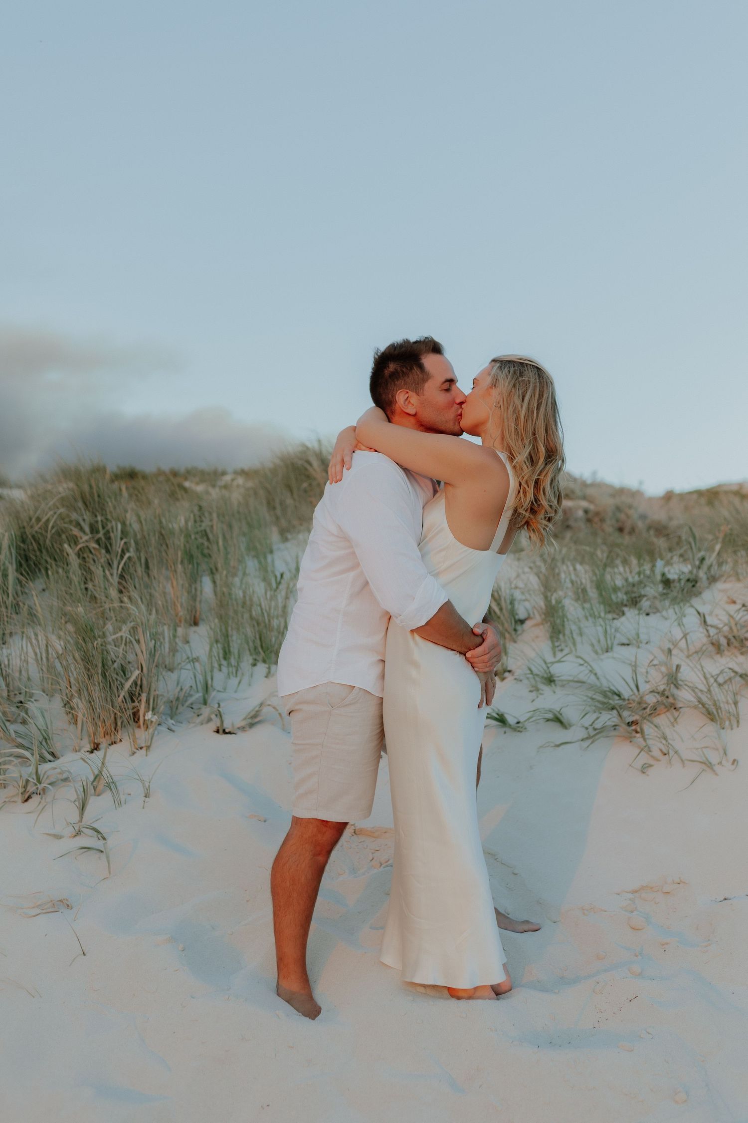 Couple kissing on white sand dunes at sunset, both wearing white outfits in a romantic beach engagement photo.