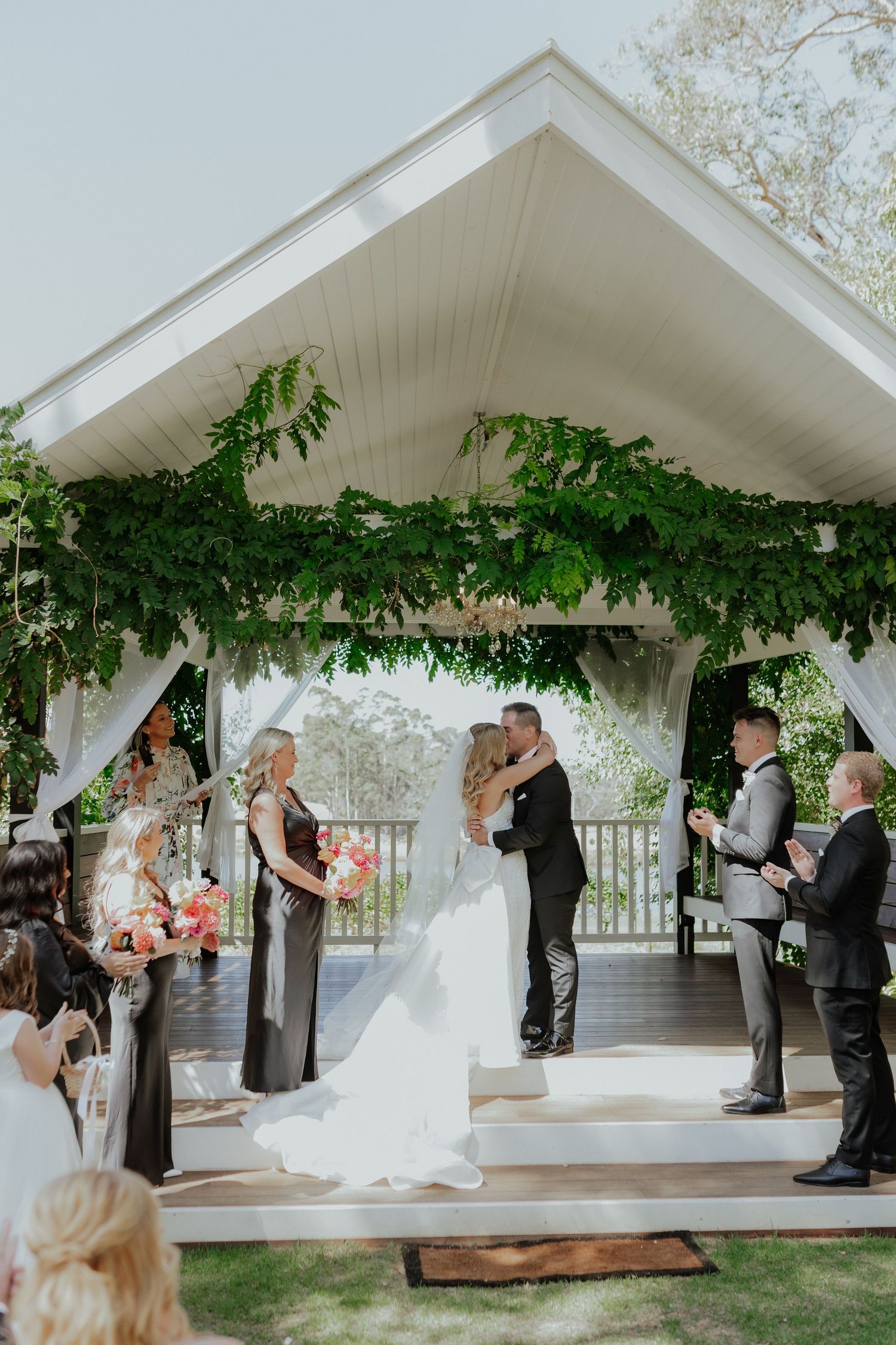 Bride and groom share first kiss under floral green arch at outdoor wedding ceremony pavilion.