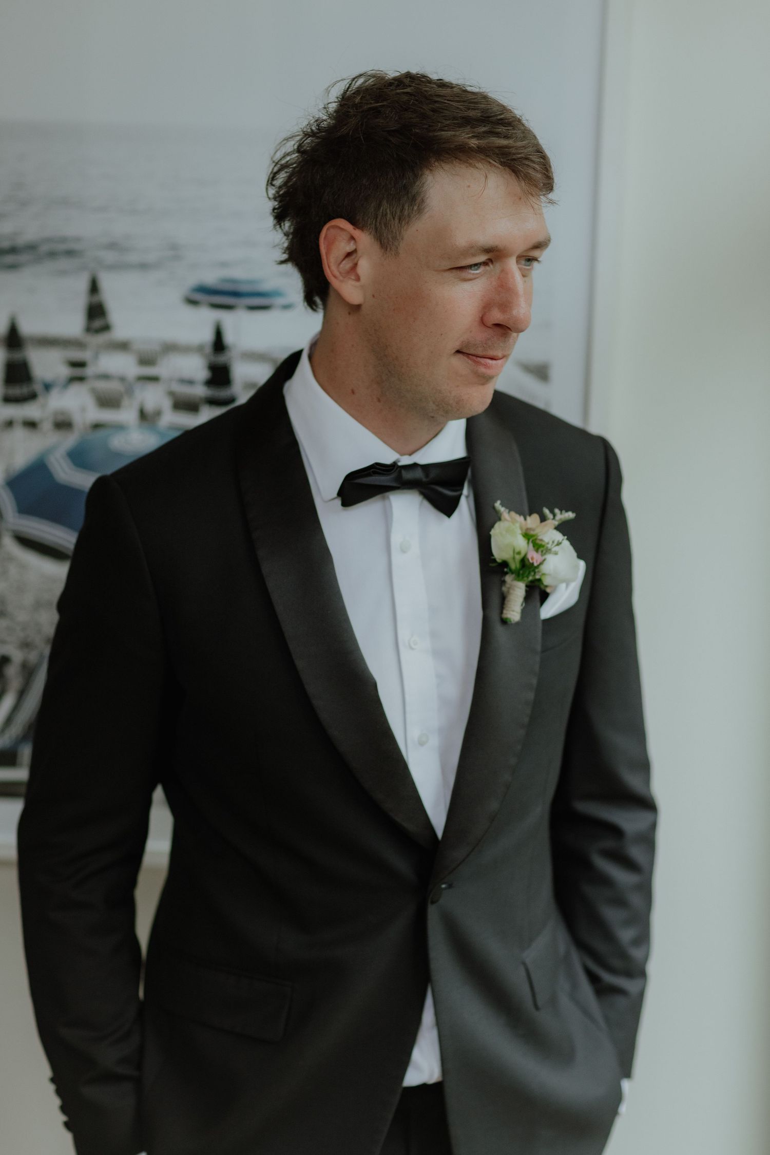 Handsome groom in black tuxedo with boutonniere gazing thoughtfully to the side indoors.