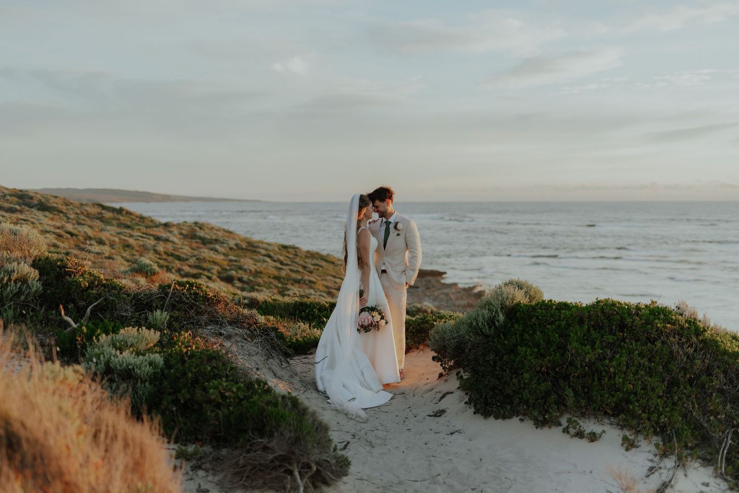 Bride and groom kissing on a sandy coastal path with ocean views at golden hour.