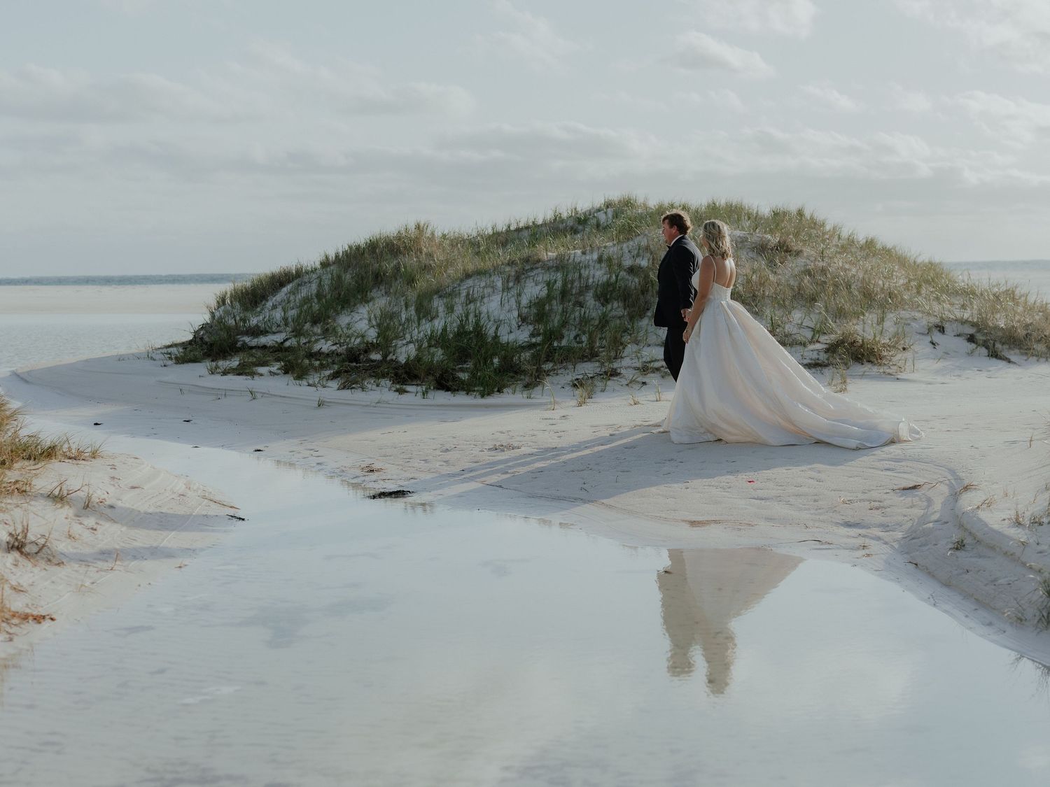 Bride and groom standing on white sand beach with reflection in shallow water at sunset.