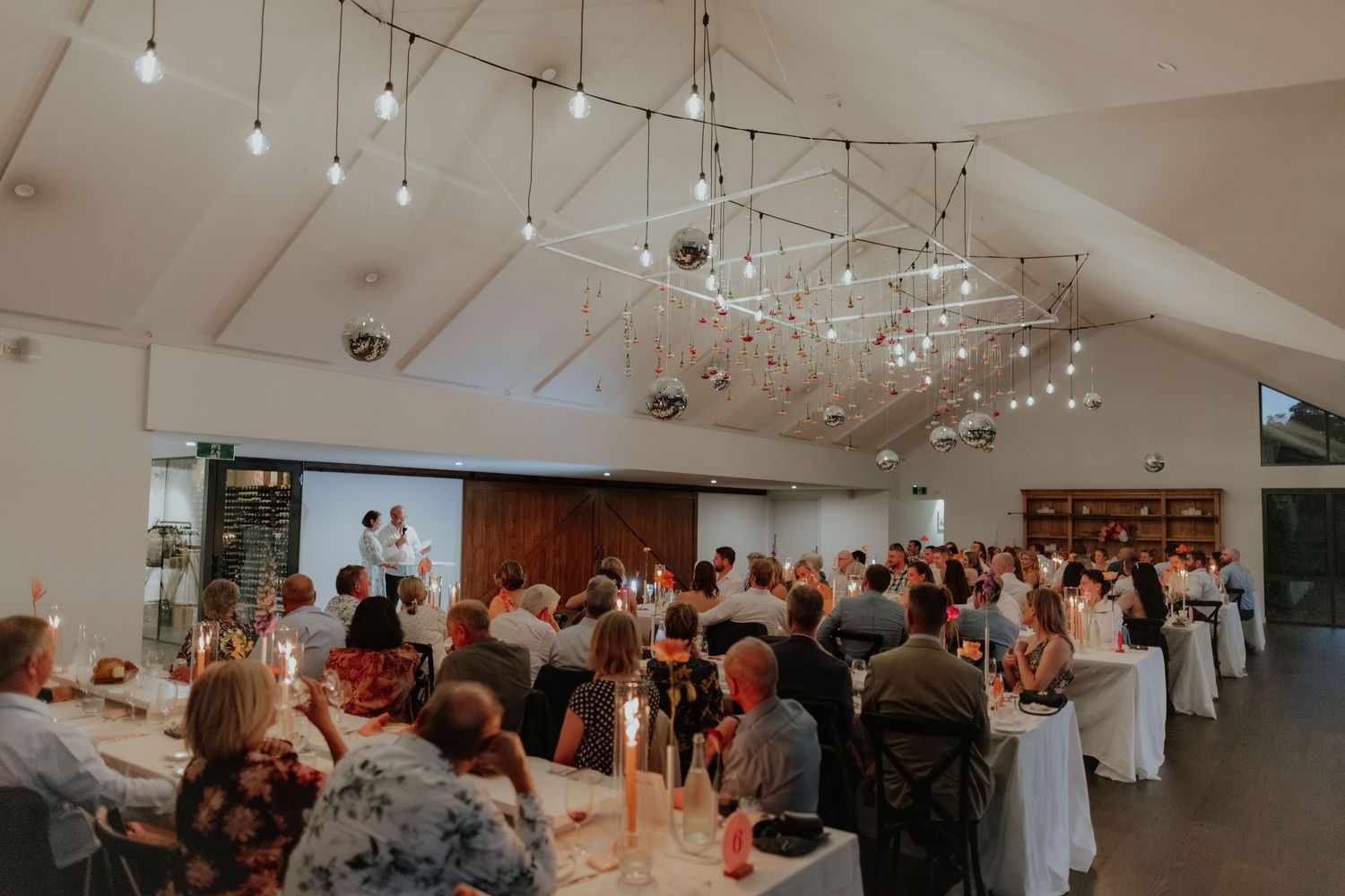 Wedding reception venue with vaulted ceiling, string lights, and guests watching a slideshow presentation.