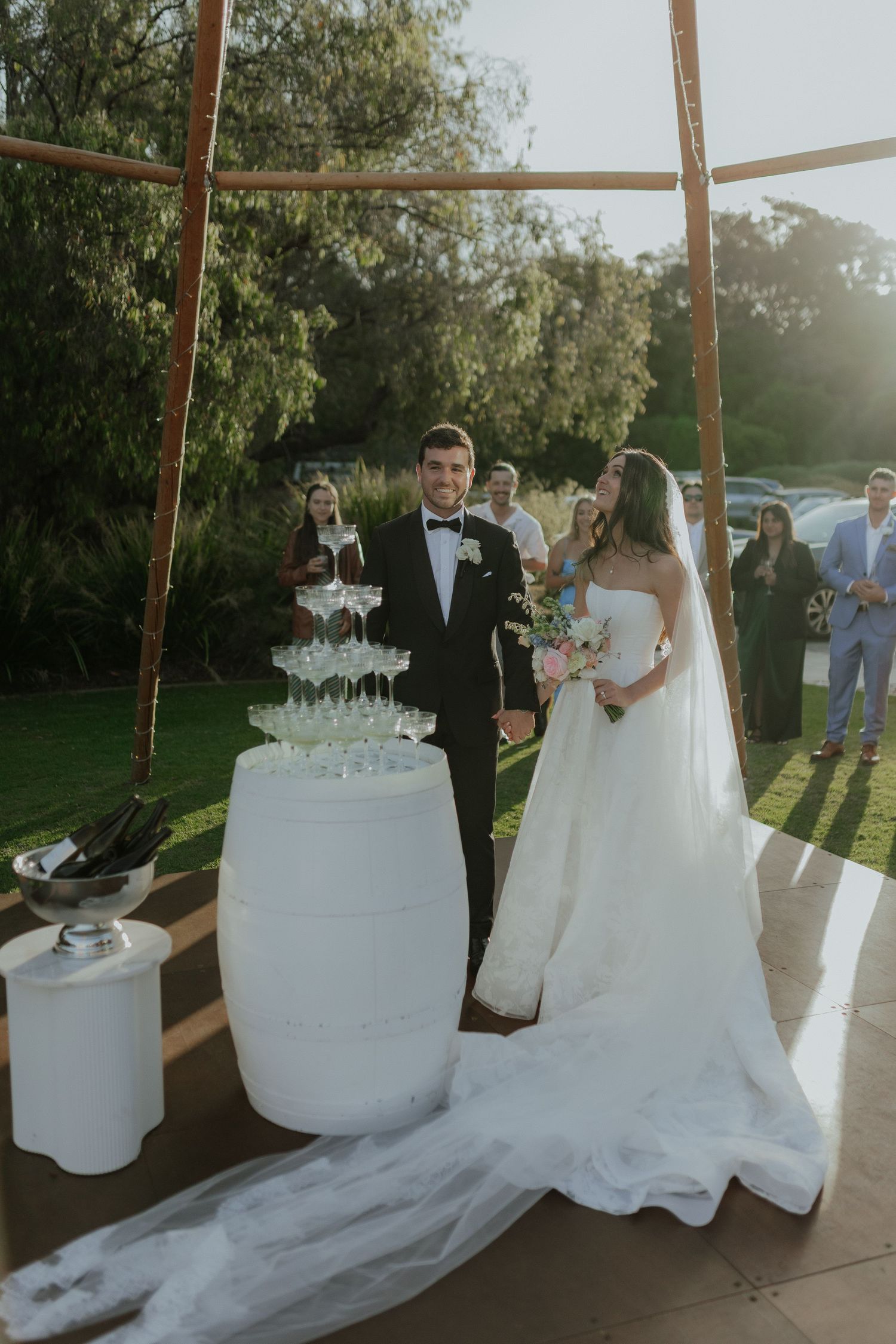 Bride and groom standing beside a champagne tower at an outdoor garden wedding reception.