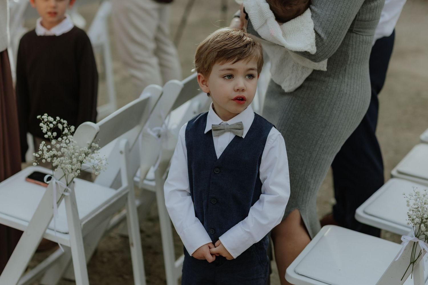 Adorable young ring bearer in navy vest and bow tie standing at outdoor wedding ceremony.