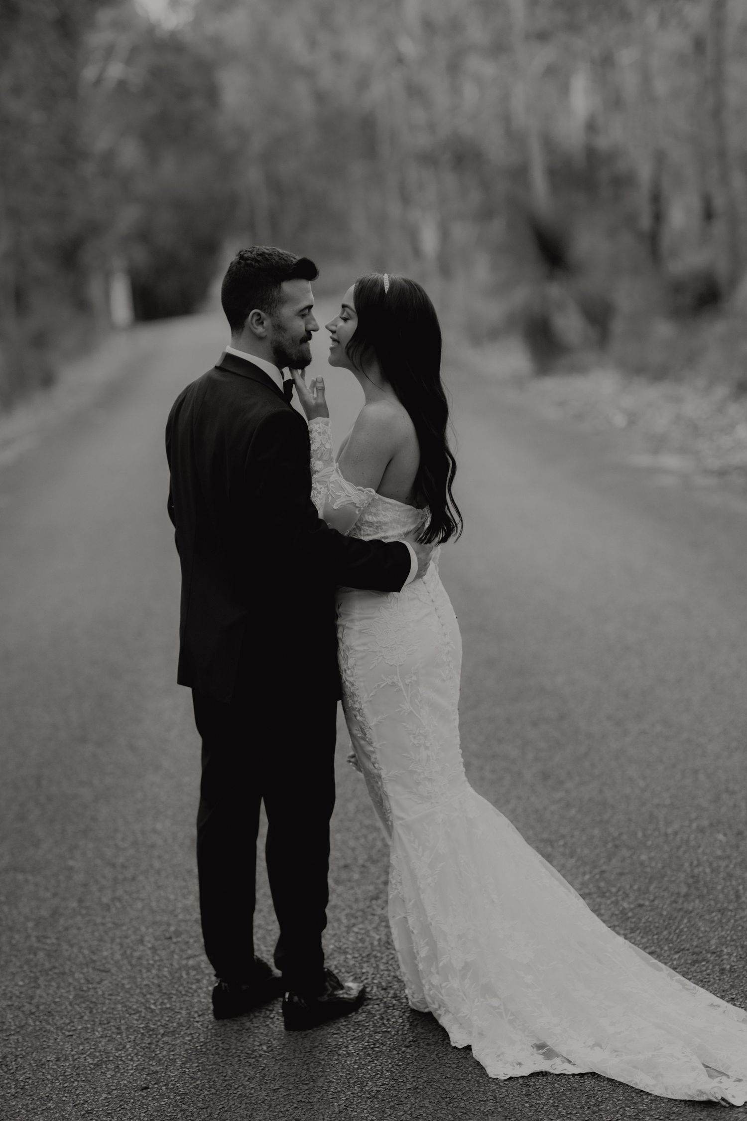 Black and white romantic portrait of bride and groom embracing on a tree-lined road.