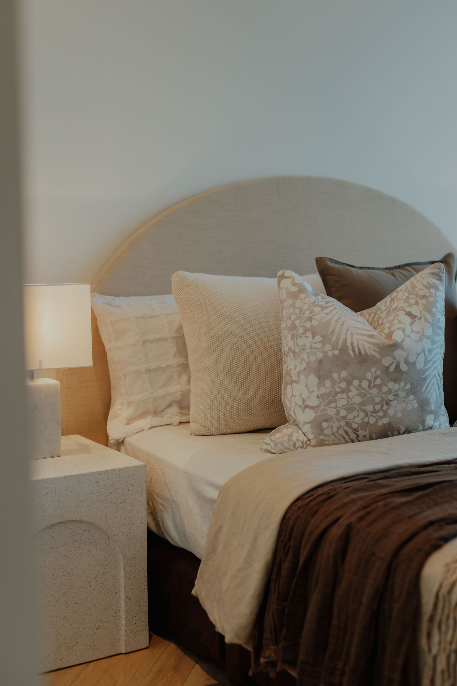 Cozy bedroom with arched upholstered headboard, layered neutral cushions, floral pillow, and warm bedside lamp.