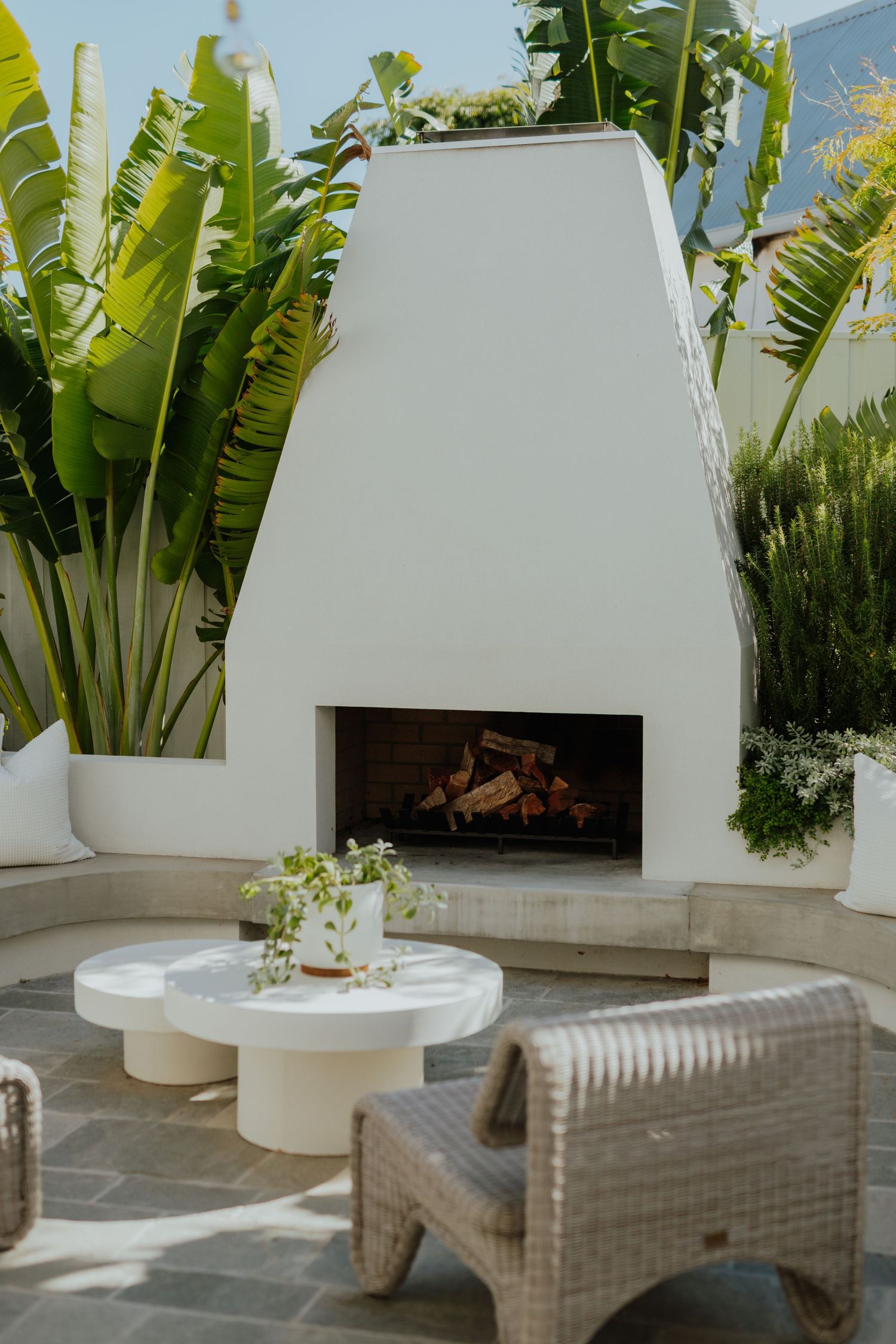 White outdoor fireplace surrounded by tropical banana palms, wicker armchair, and round concrete coffee table.