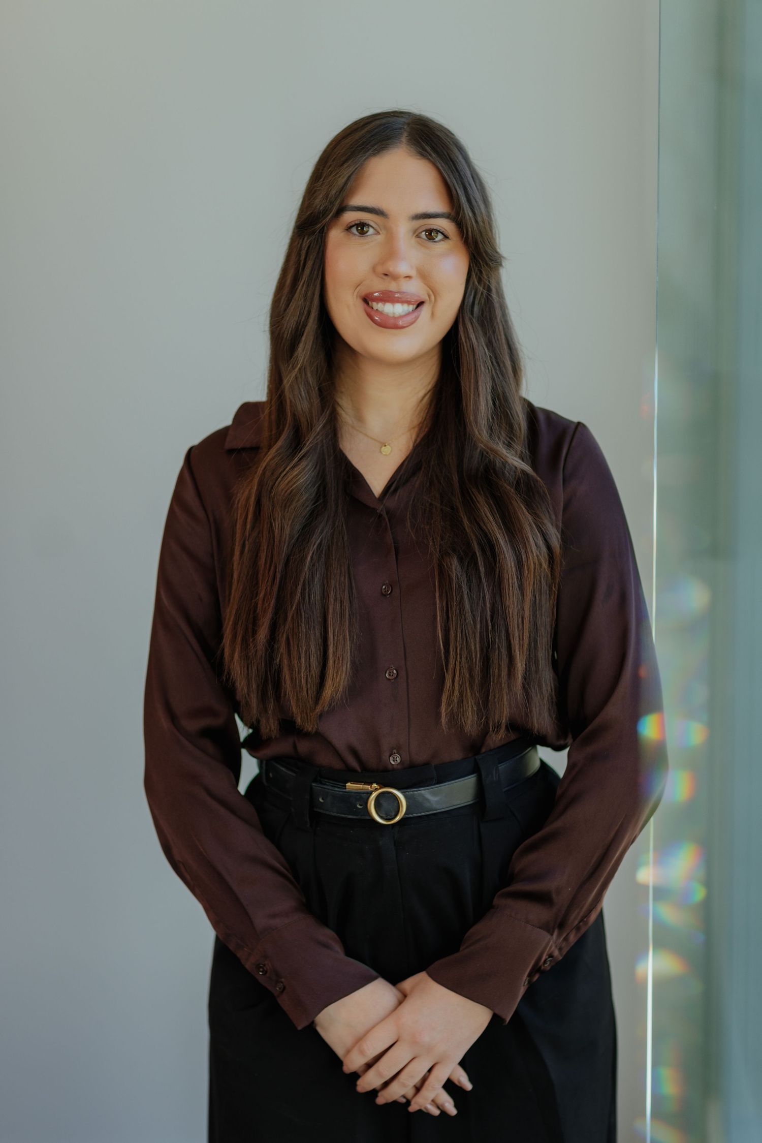 Young professional woman with long dark hair smiling in a dark burgundy blouse and black trousers.
