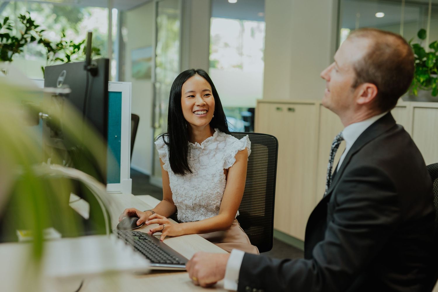 Two professionals collaborating at a desk in a modern office, woman smiling at laptop while man in suit listens.