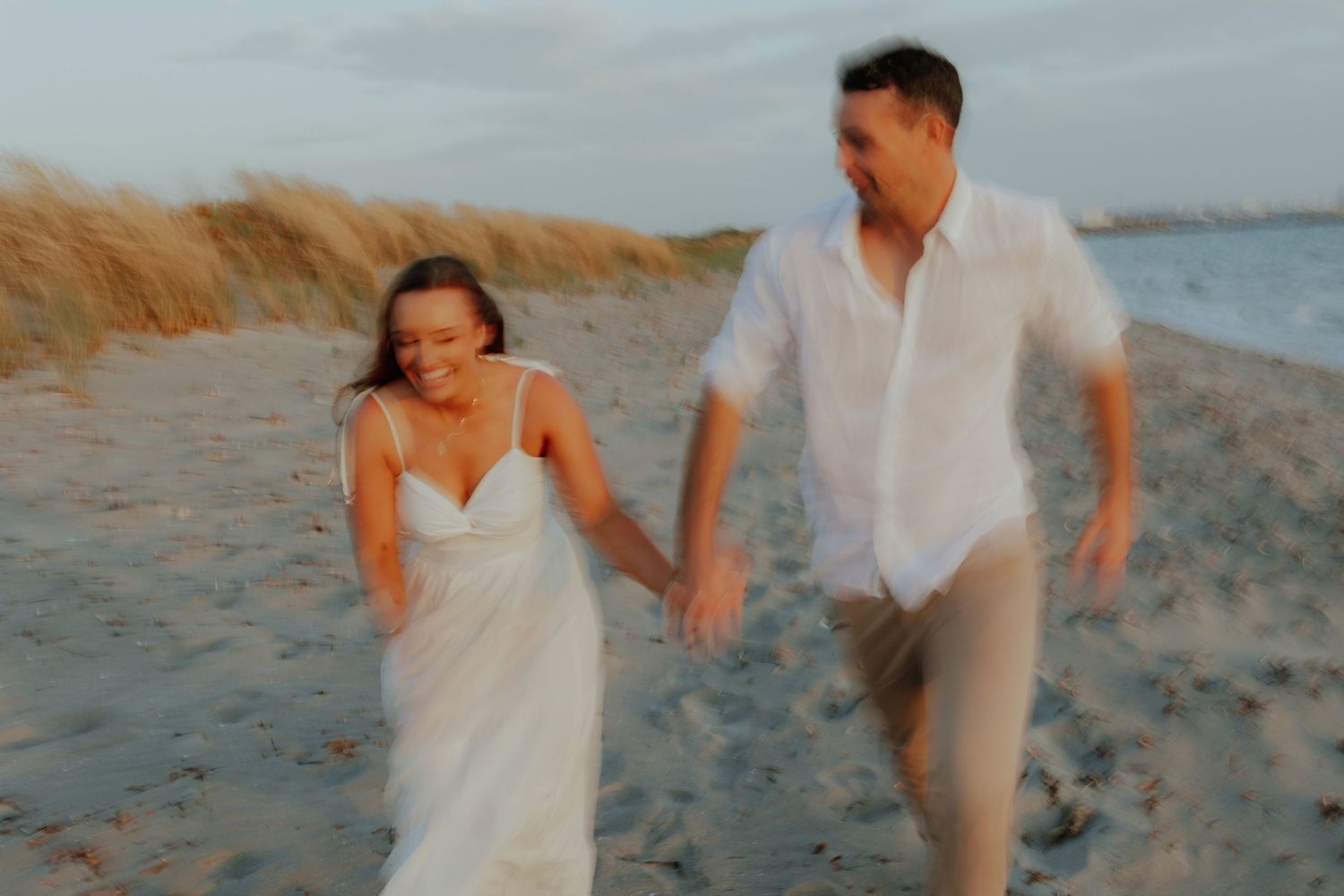 Motion-blurred couple in white holding hands running joyfully on sandy beach.
