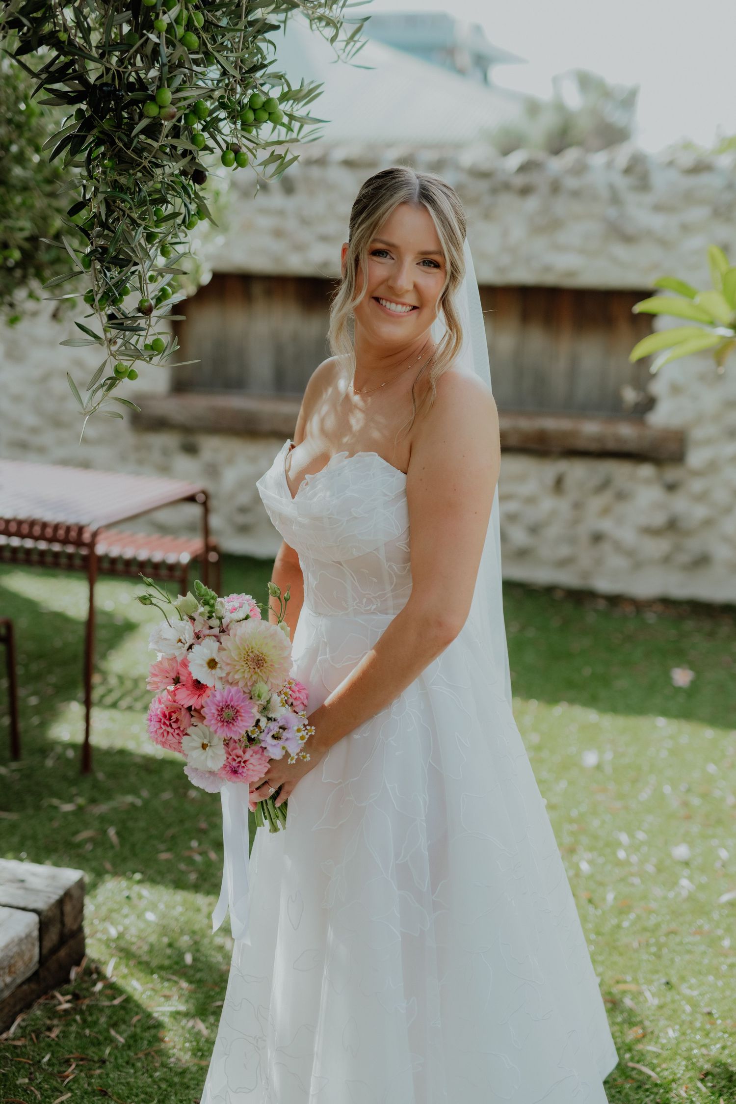 Smiling bride in strapless white lace gown holding pink floral bouquet outdoors on wedding day.