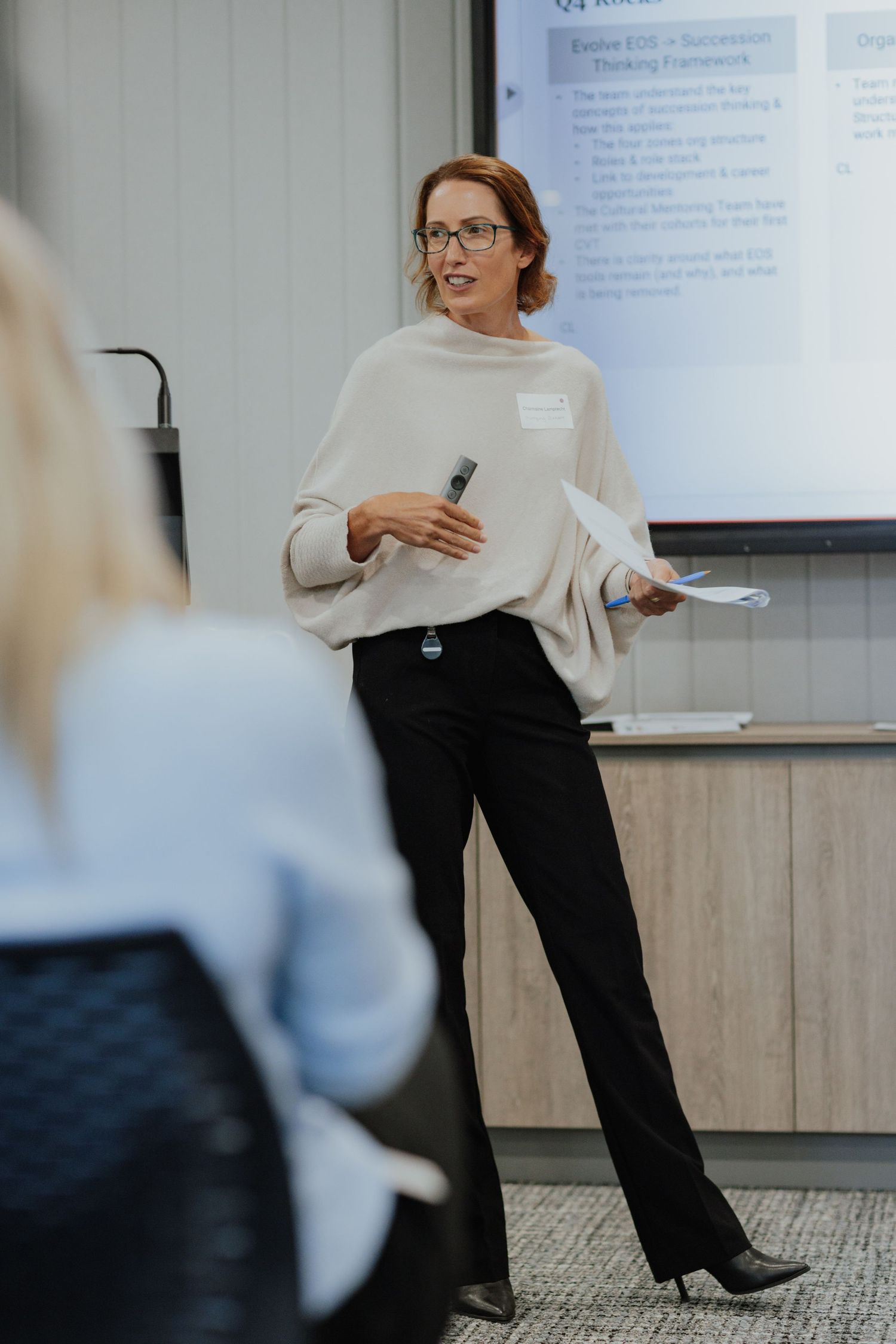 Confident woman in cream sweater and black pants presenting with a clicker at a professional business meeting.
