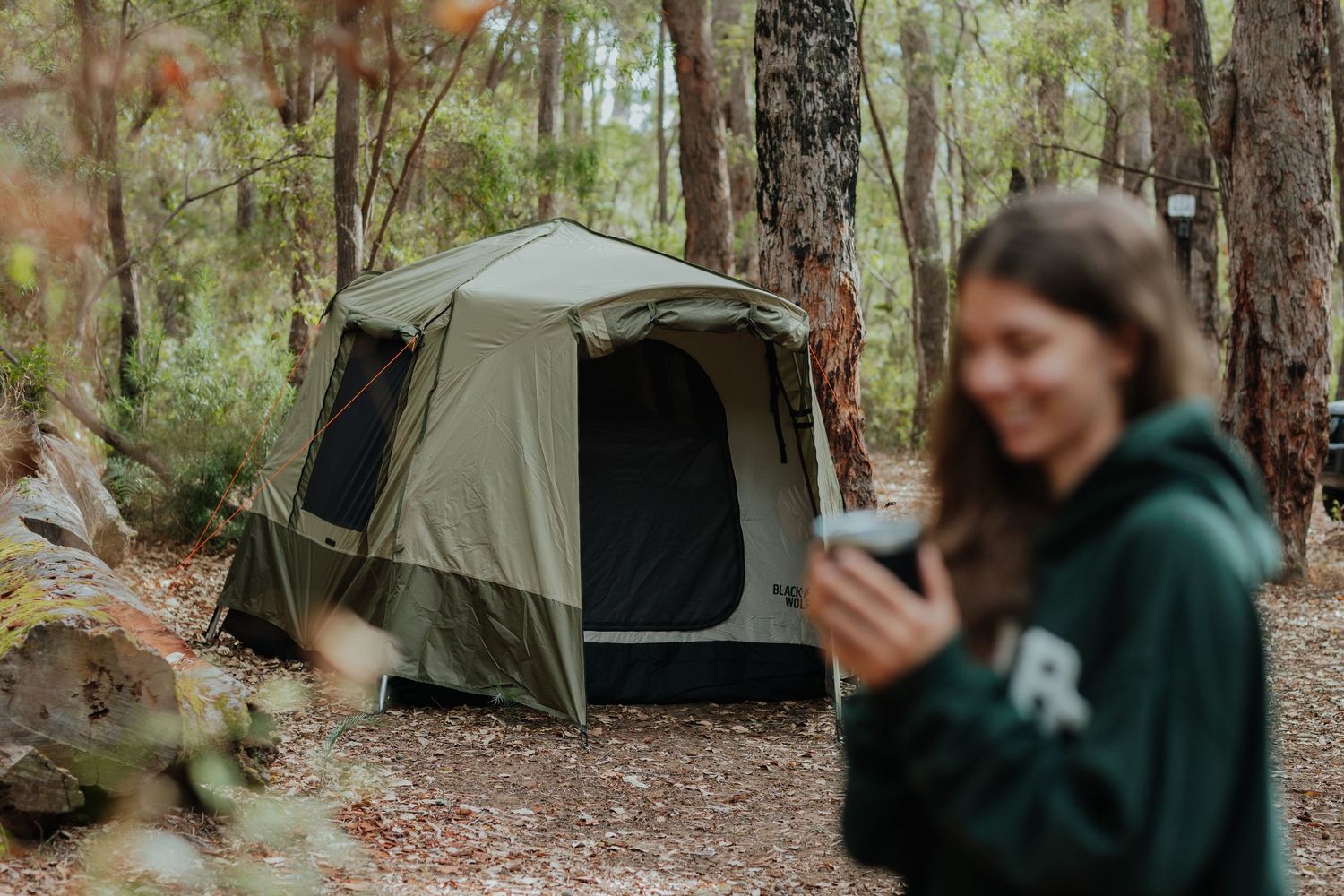 Olive green camping tent set up in a forest clearing with a blurred woman in foreground.