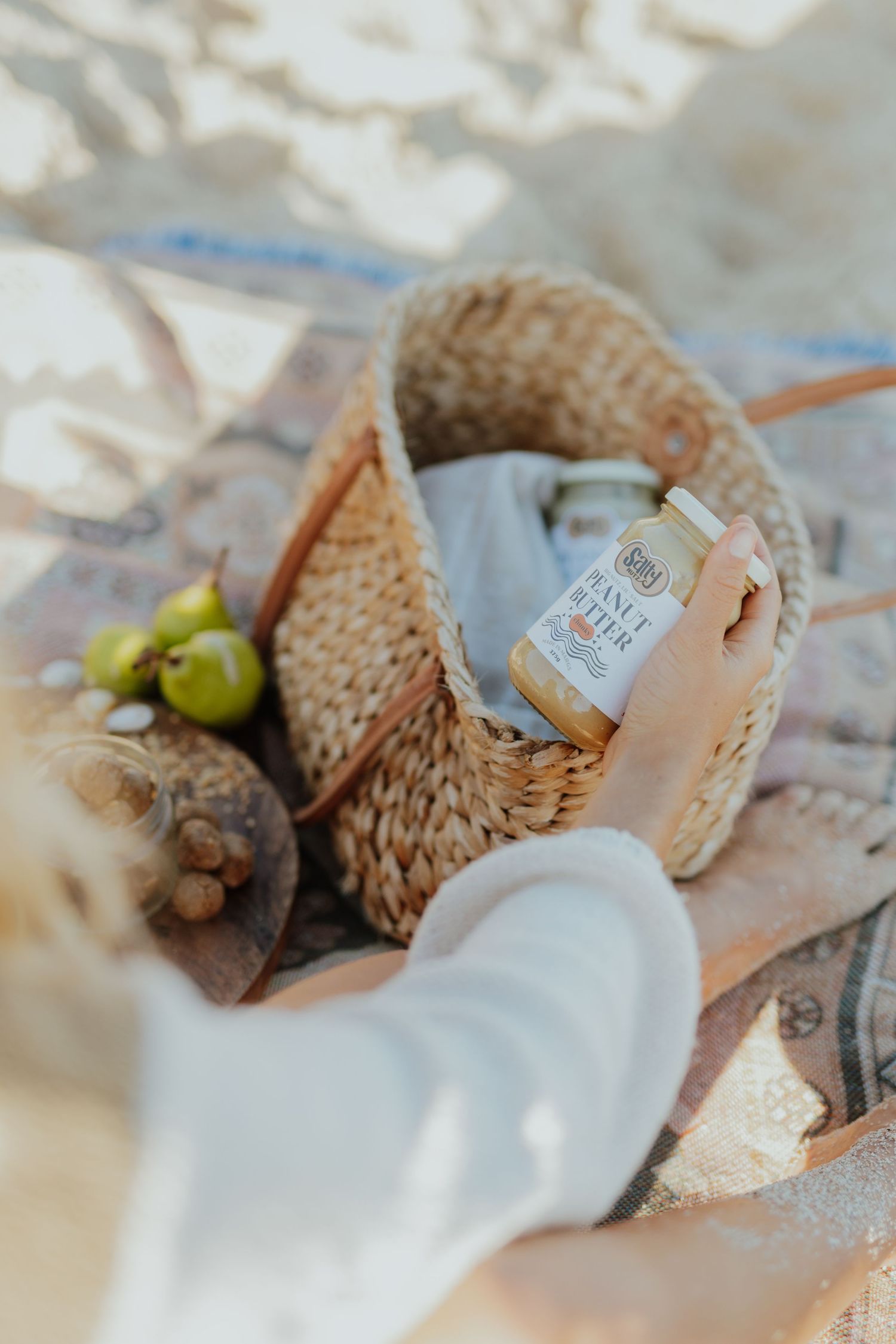 Hand placing a jar of organic nut butter into a woven straw basket with fresh fruit at a picnic.