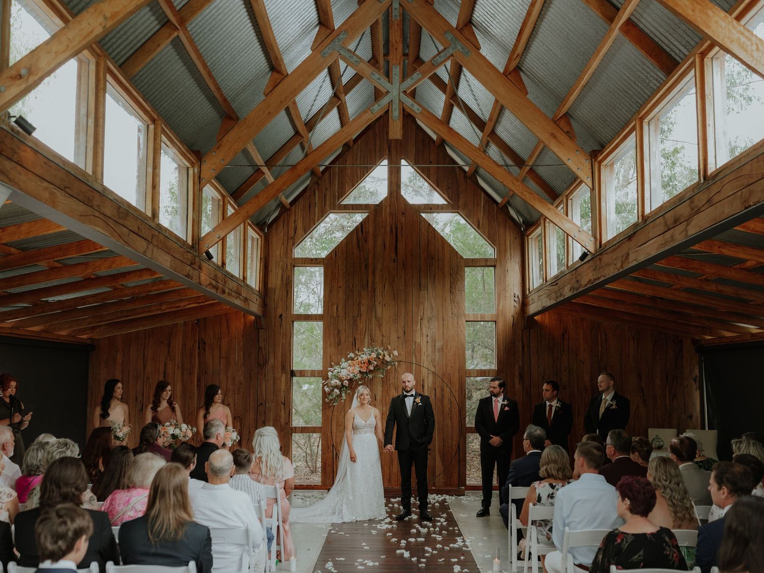 Wedding ceremony inside a rustic wooden chapel with high vaulted ceilings, guests seated in white chairs.