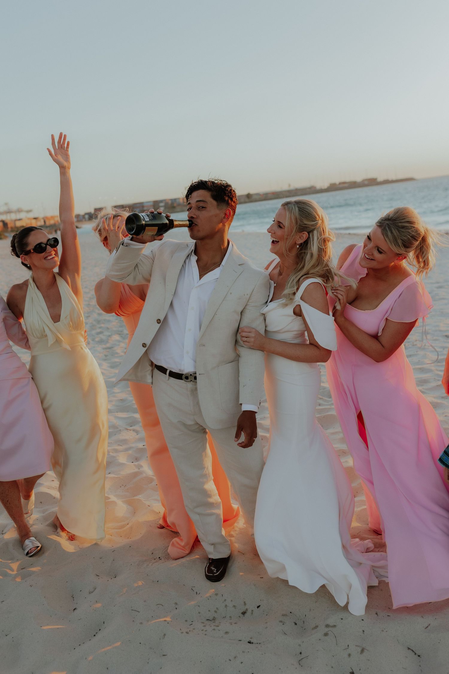 Joyful wedding party celebrating on a sandy beach at sunset with bridesmaids in pink gowns.