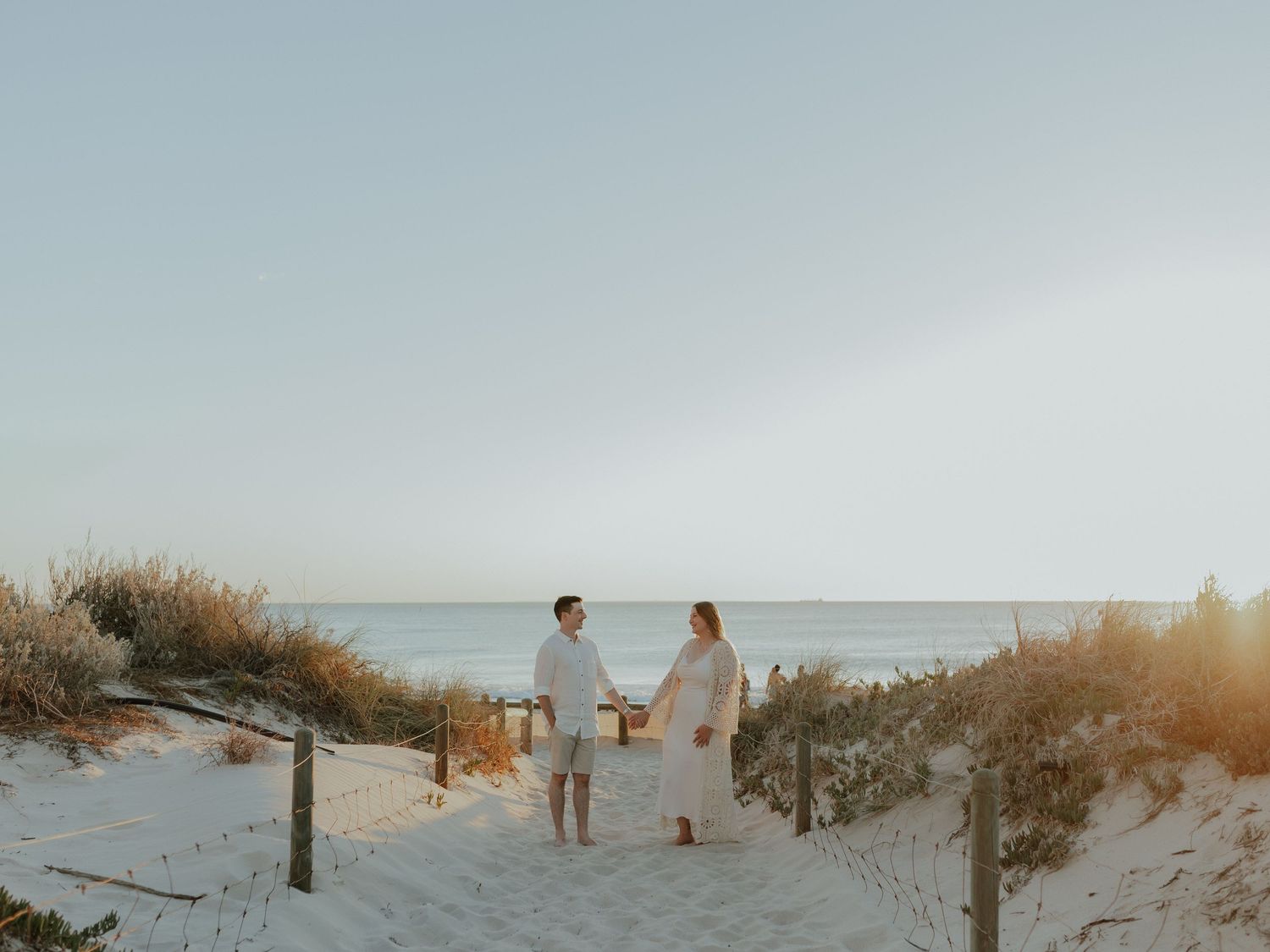 Couple in white holding hands on sandy beach path at golden sunset.