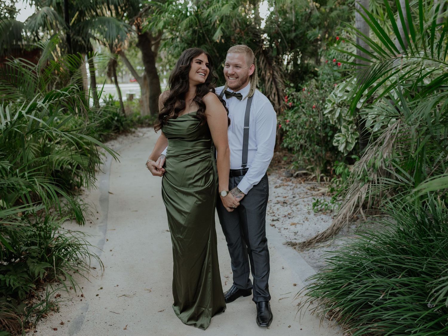 Couple smiling and holding hands on a tropical sandy path, woman wearing a satin green gown.