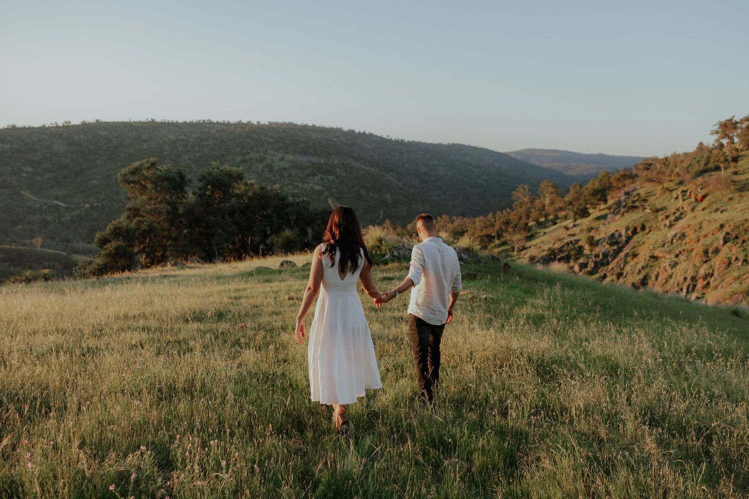 Couple holding hands walking through a golden green meadow with rolling hills in the background.