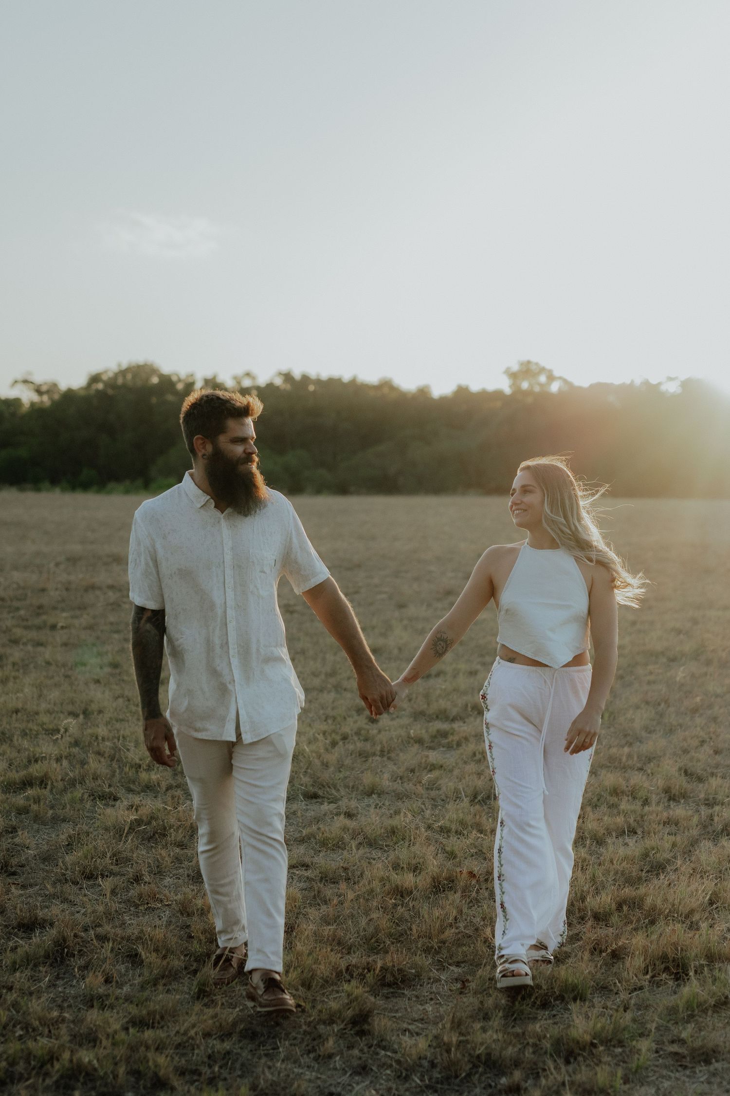 Happy couple holding hands walking through golden field at sunset, both dressed in white.