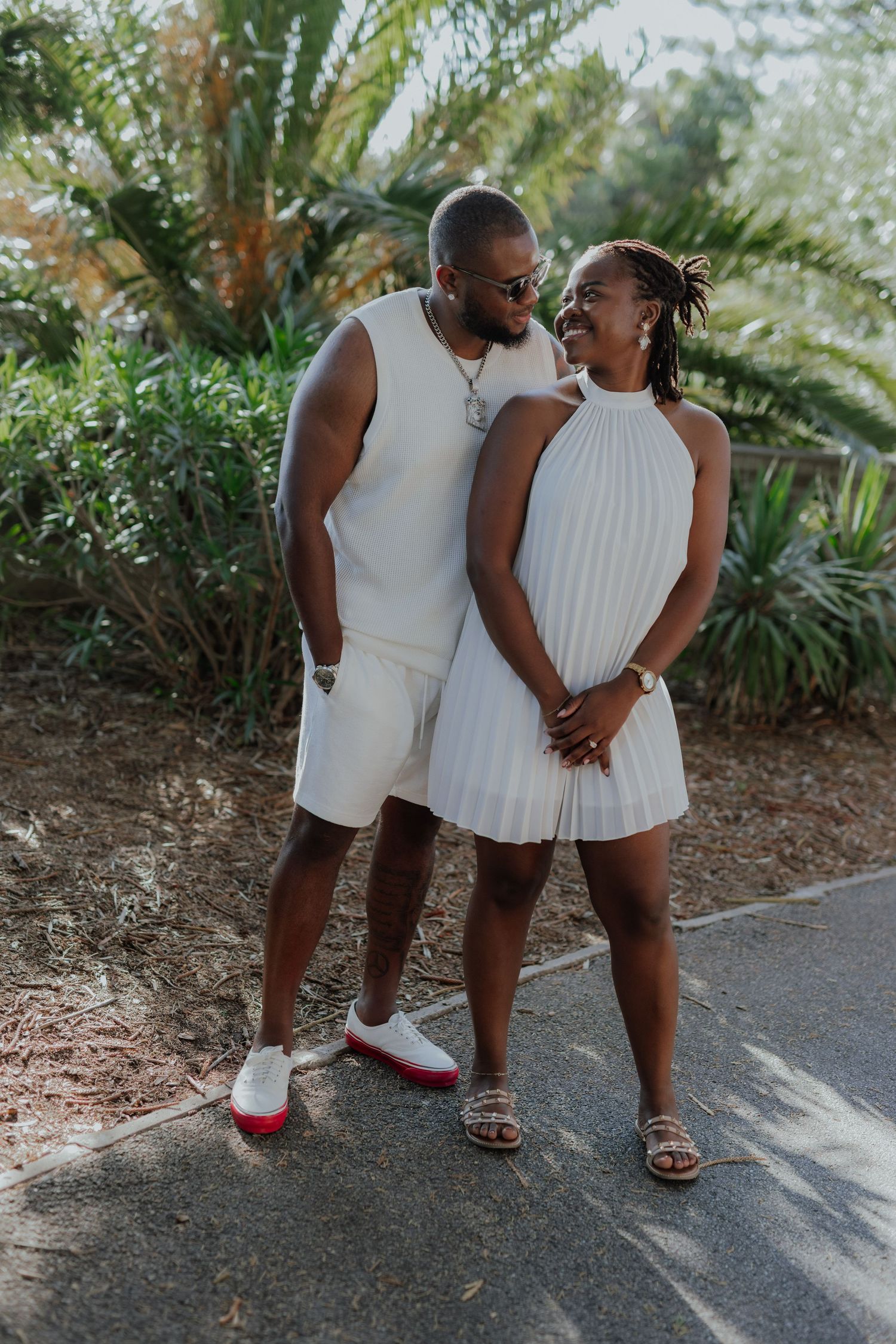 Stylish couple in matching white outfits posing together in front of lush tropical greenery.