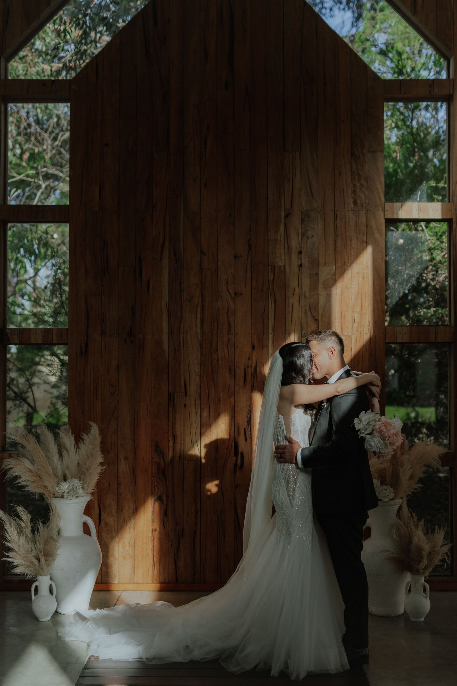 Bride and groom sharing romantic kiss in rustic wooden chapel with pampas grass decor and arched windows.