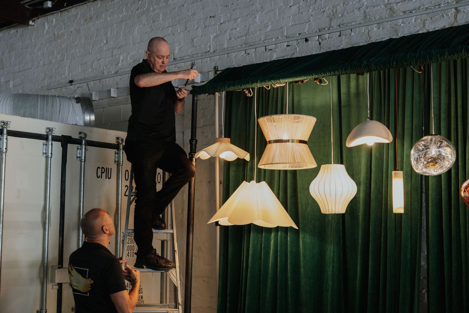 Two men installing decorative pendant lampshades on a green velvet backdrop in an event space.