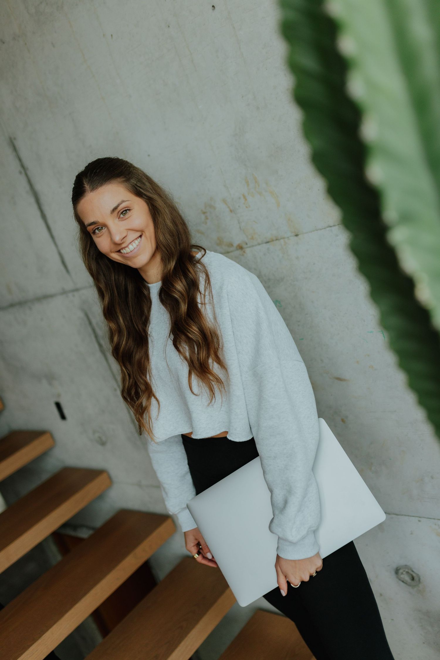 Smiling woman in grey cropped sweatshirt carrying laptop beside wooden stairs and green plant.