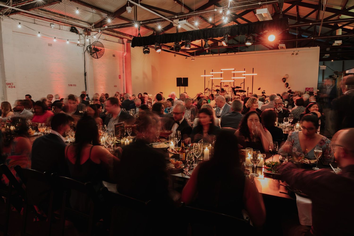 Large indoor event reception with guests dining at candlelit tables under warm string lights.